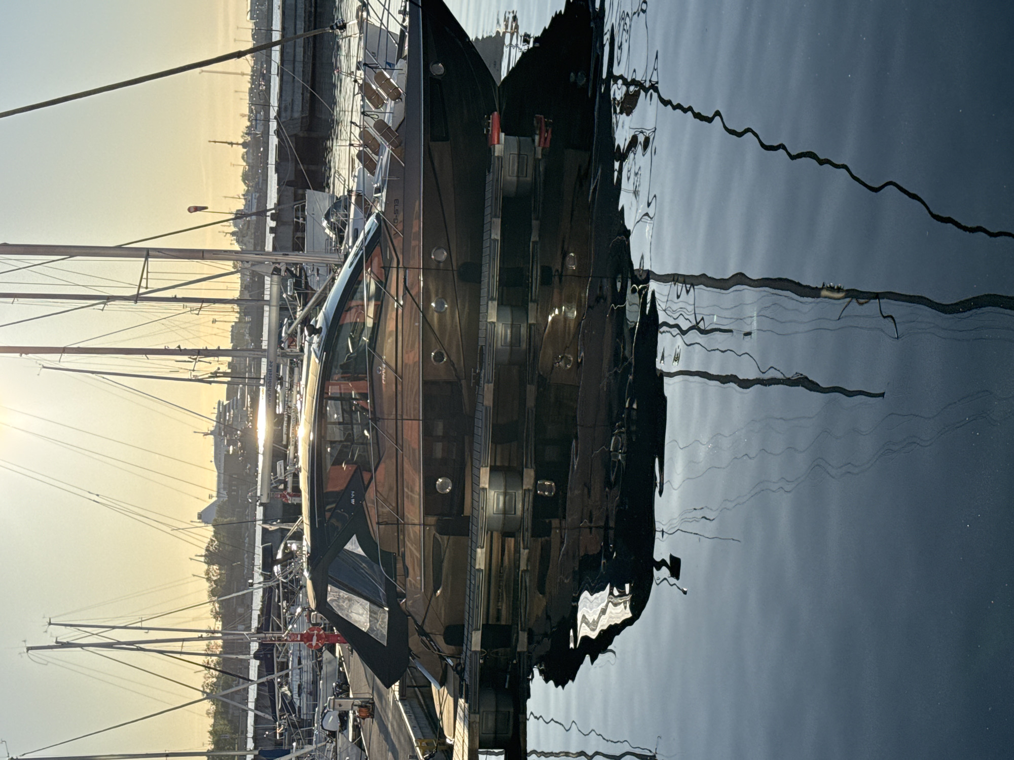 The image depicts a marina scene in Tallinn, Estonia, during either sunrise or sunset, characterized by a clear sky with a yellow-orange gradient and a bright sun causing glare in the upper center. In the foreground, a dark-hulled motor yacht, identified by the visible name "CRANCHI" and registration "0-573," is docked at a wooden pontoon. The yacht features a black canvas cover over its aft cockpit and multiple circular portholes along its hull.
The calm water surface reflects the yacht, creating a distorted mirror image, as well as the masts and rigging of other vessels. Several white sailboats with tall masts are docked in the mid-ground, and a fueling station or service point is visible on the dock to the left. A sign reading "www.hobiseil.ee" is visible on the hull of one of the boats in the mid-ground.
In the background, a large white multi-deck passenger ferry is visible, alongside a distinctive pyramidal concrete building, identifiable as Tallinn's Linnahall. Further industrial harbor infrastructure, including cranes and other ships, occupies the distant horizon. A red and white windsock is visible amongst the distant port structures. No people are present in the captured scene. The presence of the Linnahall landmark, large ferries, and the Estonian domain on the "Hobiseil" sign conclusively place the location in the port area of Tallinn, Estonia.