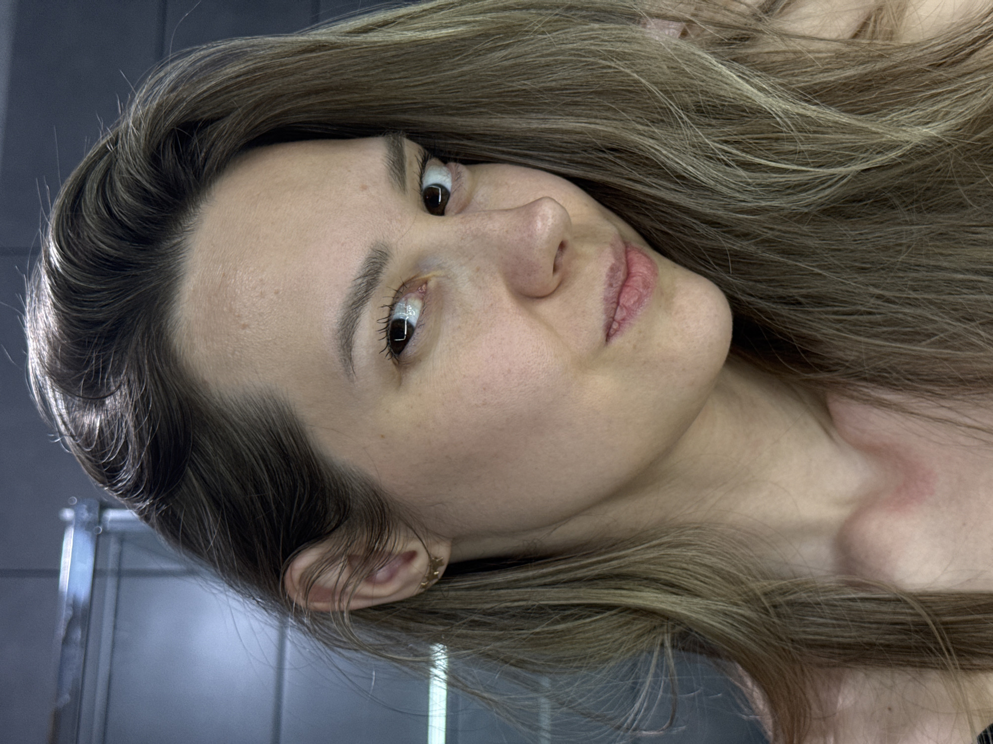 A close-up, intimate portrait features a young woman with long, light brown hair, possibly with natural highlights, framing her face. The image is oriented with her head on the left and her body extending to the right, implying she is lying down with her head tilted. Her skin is fair, revealing natural texture with visible pores and some minor blemishes or freckles. She has dark, full eyebrows and long eyelashes, with mascara visible on her lower lashes. Her dark brown eyes are wide, with her right eye looking towards the viewer and her left eye gazing slightly upwards, conveying a contemplative or curious expression. Her lips are slightly parted, showing their natural texture.
A small, intricate gold-colored earring is visible on her ear, which is partially covered by strands of her hair. Her neck and shoulder area are visible, showing a faint reddish mark on her skin. The background is dark and indistinct, featuring cool tones of gray and blue with subtle reflections and vertical elements, suggesting an indoor setting, possibly a bathroom with a glass or mirrored surface. The lighting is soft and natural, illuminating her features without harsh shadows. No specific activity is shown beyond posing for the portrait. There is no visible text in the image.