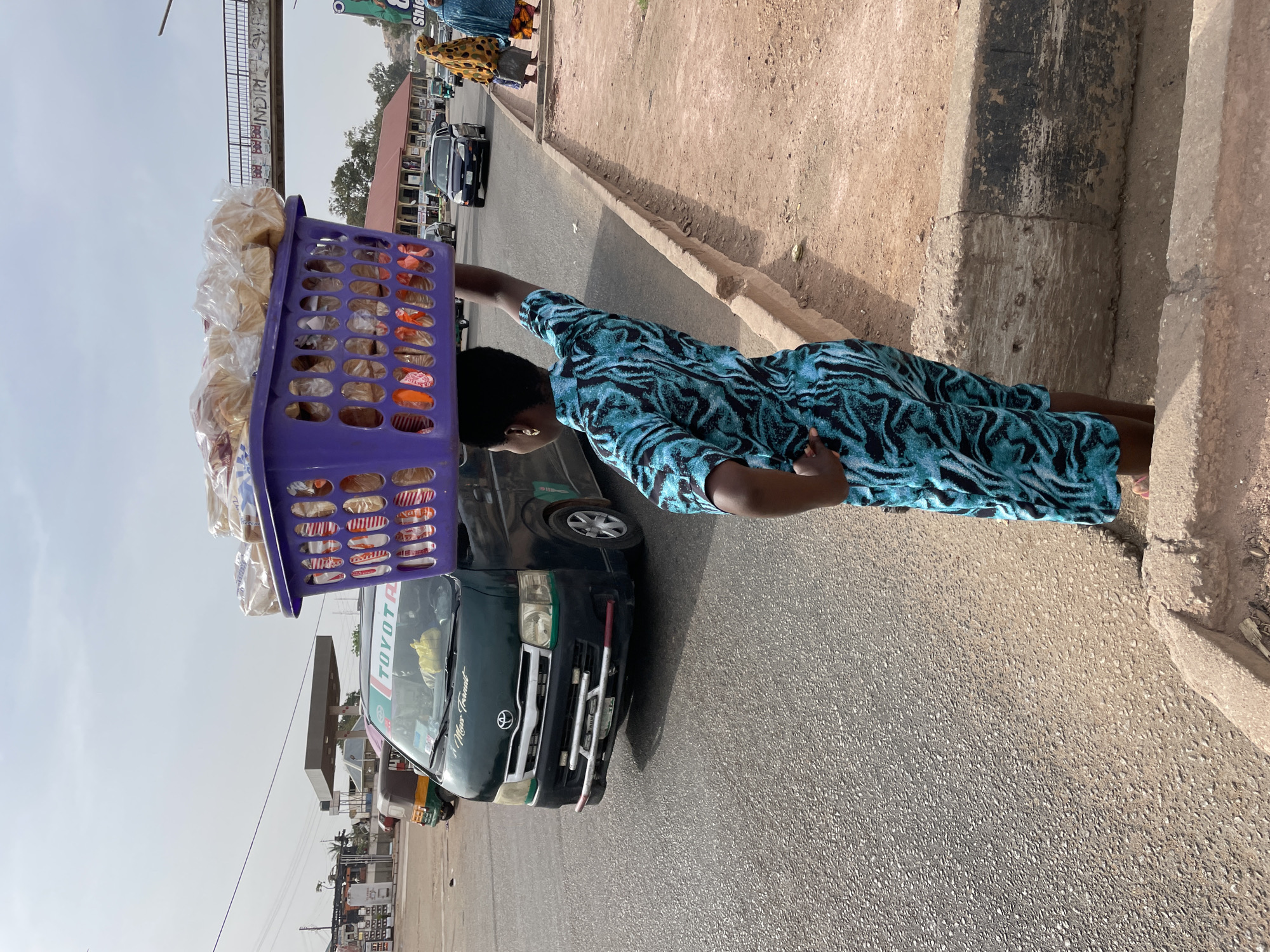 A person is captured in a street scene, carrying a large purple plastic basket filled with goods on their head. The basket primarily contains numerous loaves of bread, individually wrapped in clear plastic, and other packaged items with colorful labels. The person, viewed from behind and slightly to the side, is wearing a blue and black patterned dress. They appear to be walking alongside a paved road, possibly engaged in street vending.
To the left of the person is a dark-colored Toyota van on the road. Visible text on the van includes "TOYOTA" on its front and "A Mano Transit" below the windshield. The road itself is asphalt, and to the person's right is a dirt sidewalk bordered by a curb.
In the background, other people can be seen further down the street, along with more vehicles and buildings lining the road. Overhead, a concrete pedestrian bridge spans the street, and some graffiti is visible on its structure, including phrases like "BAD BOYS" and "INDIM". The sky is bright and clear, indicating a sunny day, likely midday or afternoon, with good visibility. The overall setting is a bustling urban or semi-urban environment.