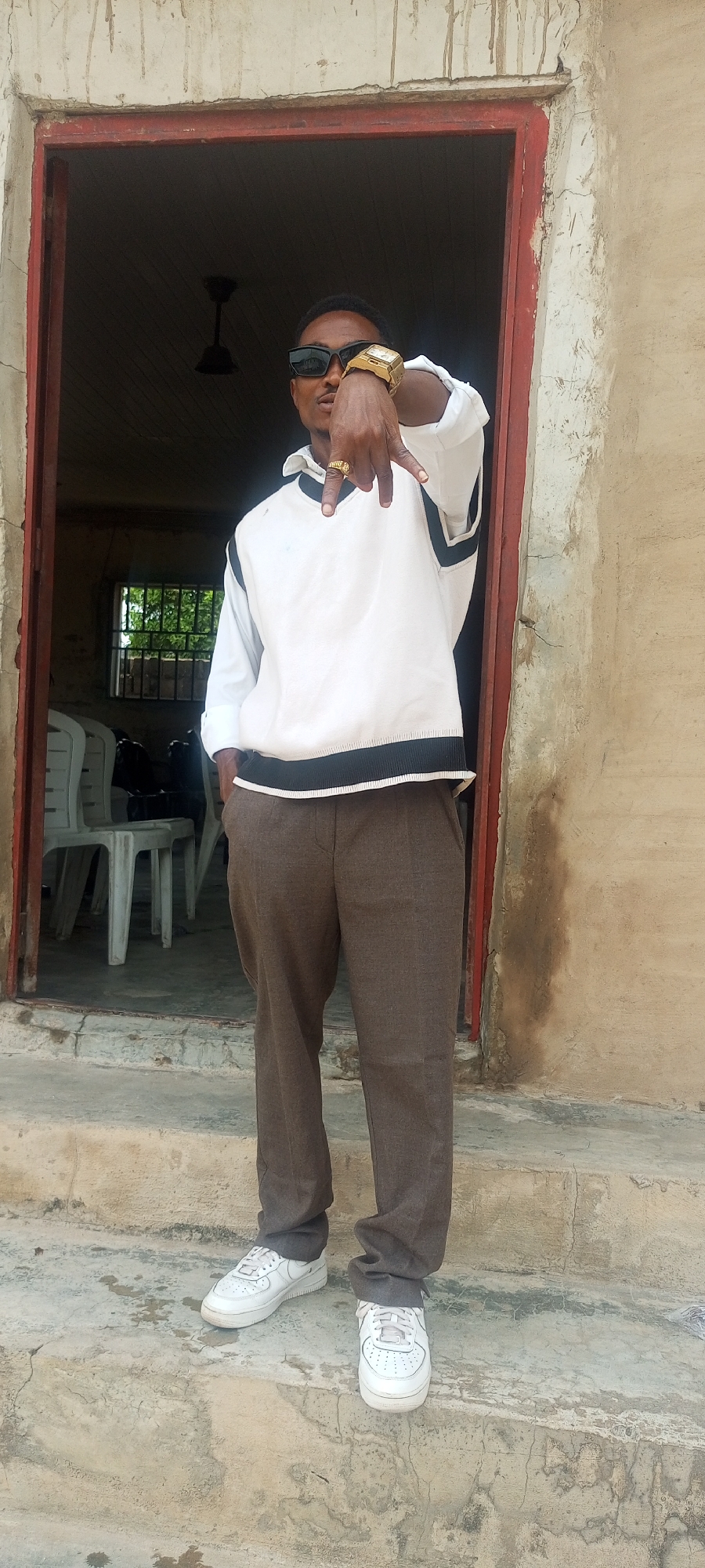 A man stands in the doorway of a building, likely in Lokoja, Nigeria, as suggested by the setting. He is wearing sunglasses, a gold watch, a white shirt with a black-trimmed sweater vest, brown trousers, and white sneakers. He is making a hand gesture, extending his index and pinky fingers while holding the others down with his thumb. Behind him, inside the building, are plastic chairs and a window. The doorway is framed in red, and the building appears to be made of concrete. The floor in front of the door consists of concrete steps.
