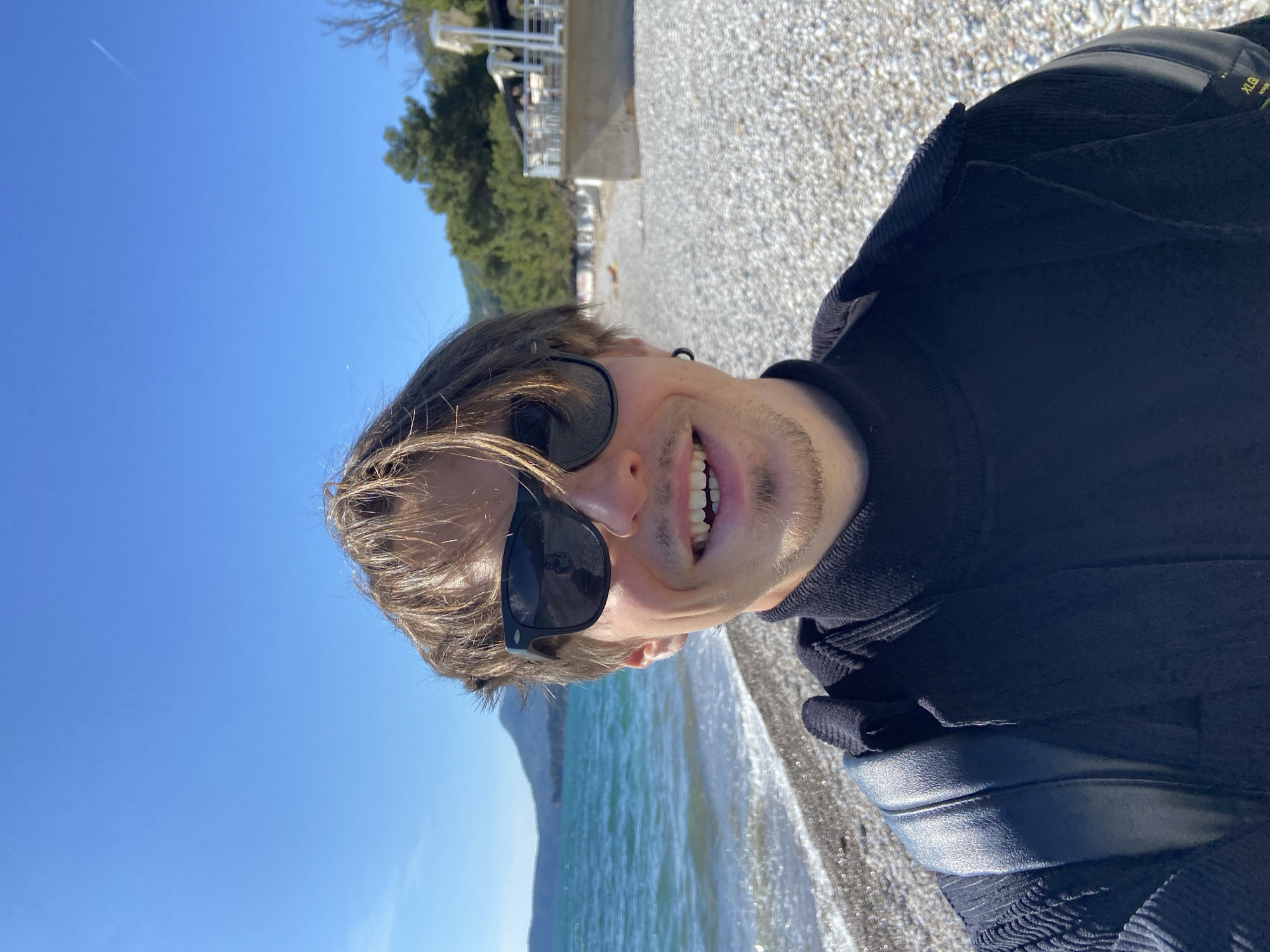 A selfie taken on a beach in Bar, Montenegro. A man with light brown hair and sunglasses smiles at the camera. He is wearing a black turtleneck and has a black bag slung over his shoulder. The beach is composed of small pebbles. The Adriatic Sea is in the background, with a mountain visible in the distance. The sky is blue and clear. Buildings are partially visible in the background, to the left.