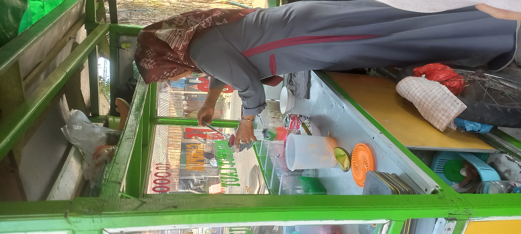 In Melayu, Indonesia, a woman wearing a maroon hijab and a grey tunic is preparing a drink from a street food cart. The cart has a green metal frame and glass windows displaying a price of "00001". Various ingredients are visible in clear plastic containers, including green jelly, colorful layered drinks, and diced fruit. The woman is holding a ladle over a transparent container, as if serving or mixing a beverage. On the counter, there is an avocado, an orange lid, and a can of Nestle's Carnation milk. A blue plastic basket filled with utensils is stored below the counter, alongside other items like plastic bags and a checkered cloth. The background suggests an outdoor setting with buildings and trees.