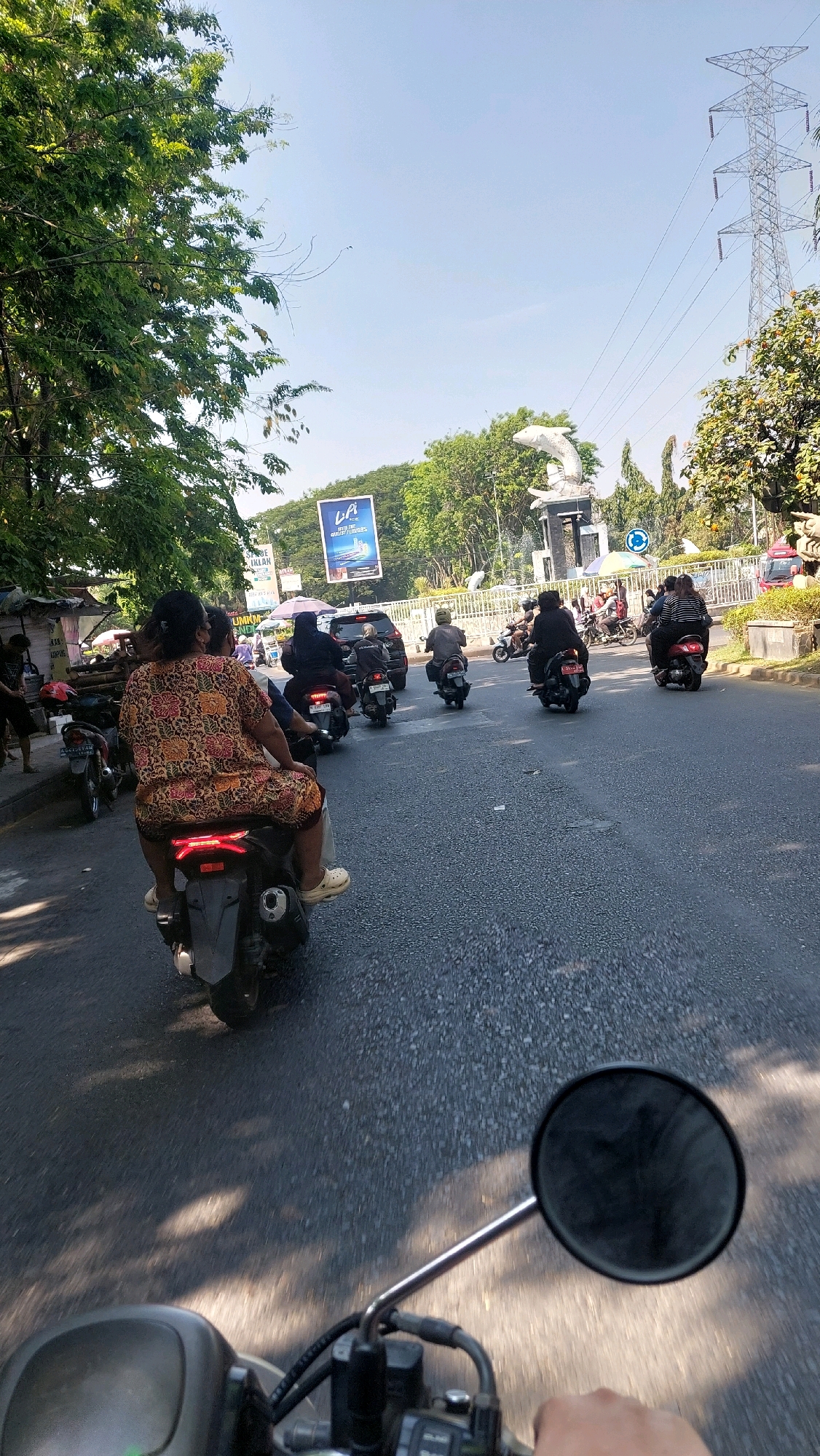 The image shows a street scene in Sepande, Indonesia, on a sunny day. The perspective is from a motorcycle, looking forward and slightly to the right. A woman wearing a brightly patterned dress is riding on the back of a scooter in the foreground, facing away from the camera. She is wearing white clogs. Several other scooters, some with multiple riders, are traveling on the asphalt road. In the background, there are trees, a large pylon, and a statue of two dolphins. A billboard advertising "LA" is also visible. The road appears to be a main thoroughfare with some traffic. The overall impression is of a typical day in a bustling Indonesian city.