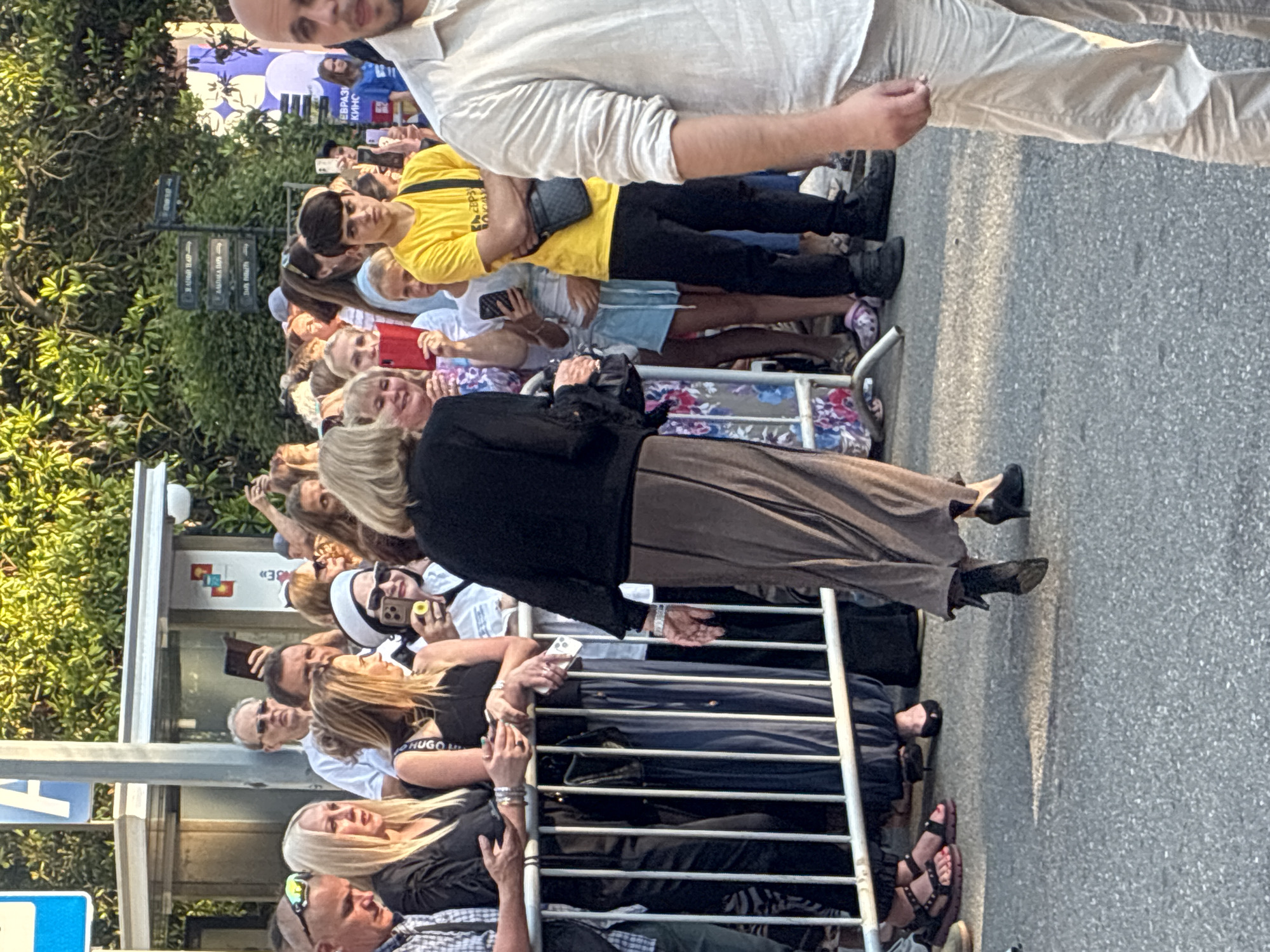 The image captures a crowd of people, likely attending an outdoor event in Sochi, Russia. The scene is oriented vertically, with the subjects and background appearing sideways. In the foreground, a woman wearing a black top and a long, flowing brown skirt is visible, along with other individuals in casual attire. Many people are holding up their phones, suggesting they are either taking pictures or videos of something or someone out of frame. A metal barricade runs horizontally across the image, separating the crowd from an unseen area, possibly a red carpet or a stage. Behind the crowd, lush green trees and a street with a paved surface are visible.
In the background, several street signs are present, though the text is not entirely legible. One sign appears to have Cyrillic lettering. The time of day seems to be late afternoon or early evening, given the warm, soft lighting and the long shadows cast on the ground. The weather appears to be pleasant, conducive to an outdoor gathering. The overall mood is one of anticipation and excitement, as indicated by the engaged expressions and the widespread use of mobile phones for recording. The presence of a banner with colorful graphics and text in the background suggests a public event or festival.