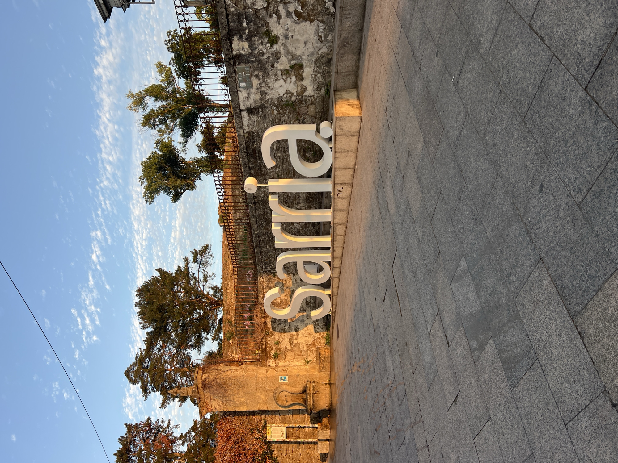 A wide-angle, eye-level photograph captures a large white sculpture spelling out "Sarria" in front of a weathered stone wall in Sarria, Spain. The sculpture is positioned on a raised concrete platform. Behind the sculpture, the stone wall exhibits signs of age with some crumbling sections and is adorned with a black wrought-iron fence at the top. Several trees with lush green foliage are visible behind the fence. The sky above is a clear blue with scattered, wispy white clouds. To the left of the sculpture, a stone structure resembling a fountain or monument stands next to a textured stone wall. The ground in the foreground is paved with large, grey granite tiles. The lighting suggests it is either early morning or late afternoon, casting warm, golden light across the scene. There are no people visible in the image.