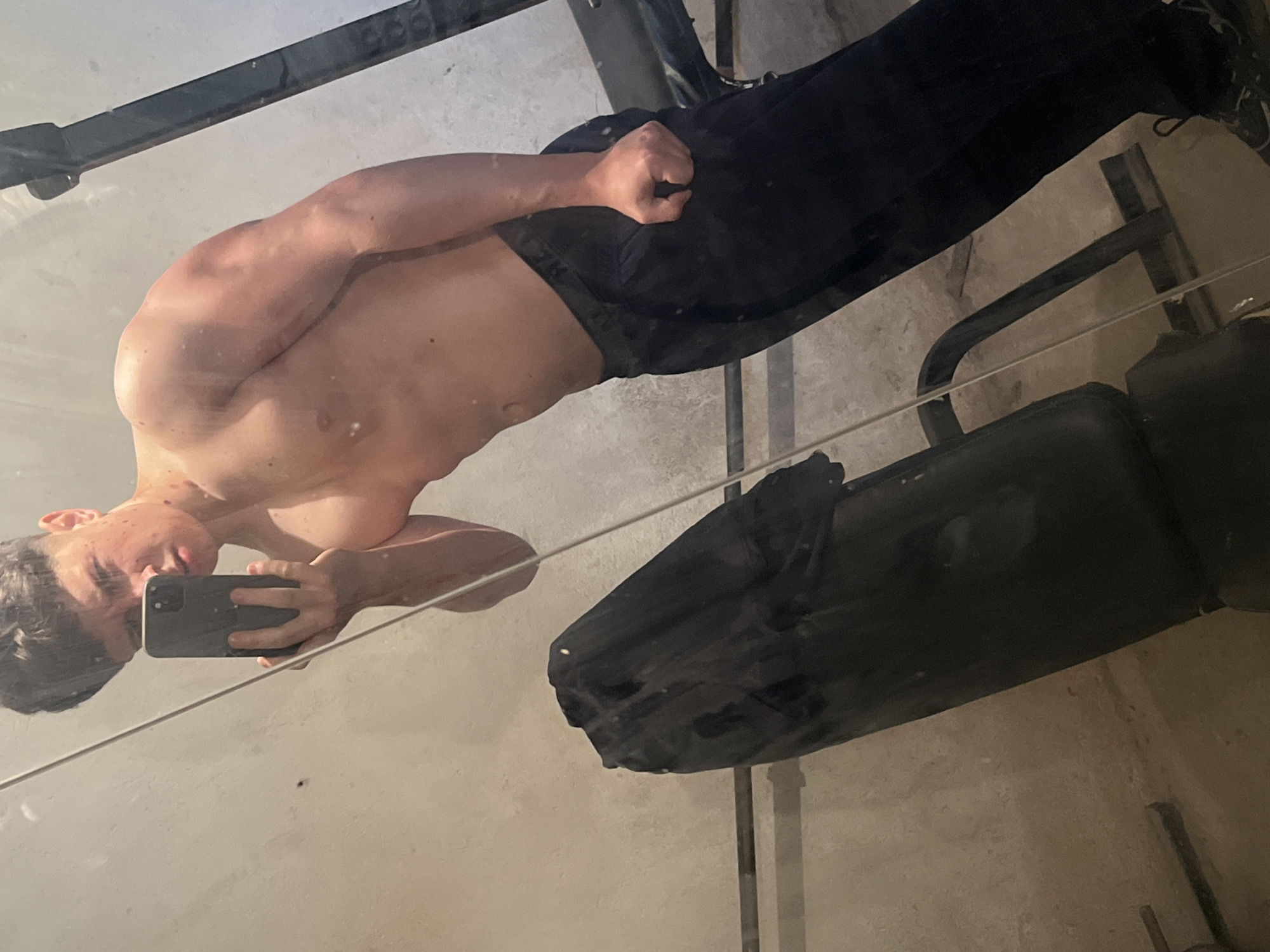 A shirtless male stands at an angle, holding a smartphone, presumably taking a selfie in a mirror. The reflection shows his upper body with visible musculature. He is wearing dark pants. The background shows gym equipment, including what appears to be a weight bench. The location is not discernible from the image.
