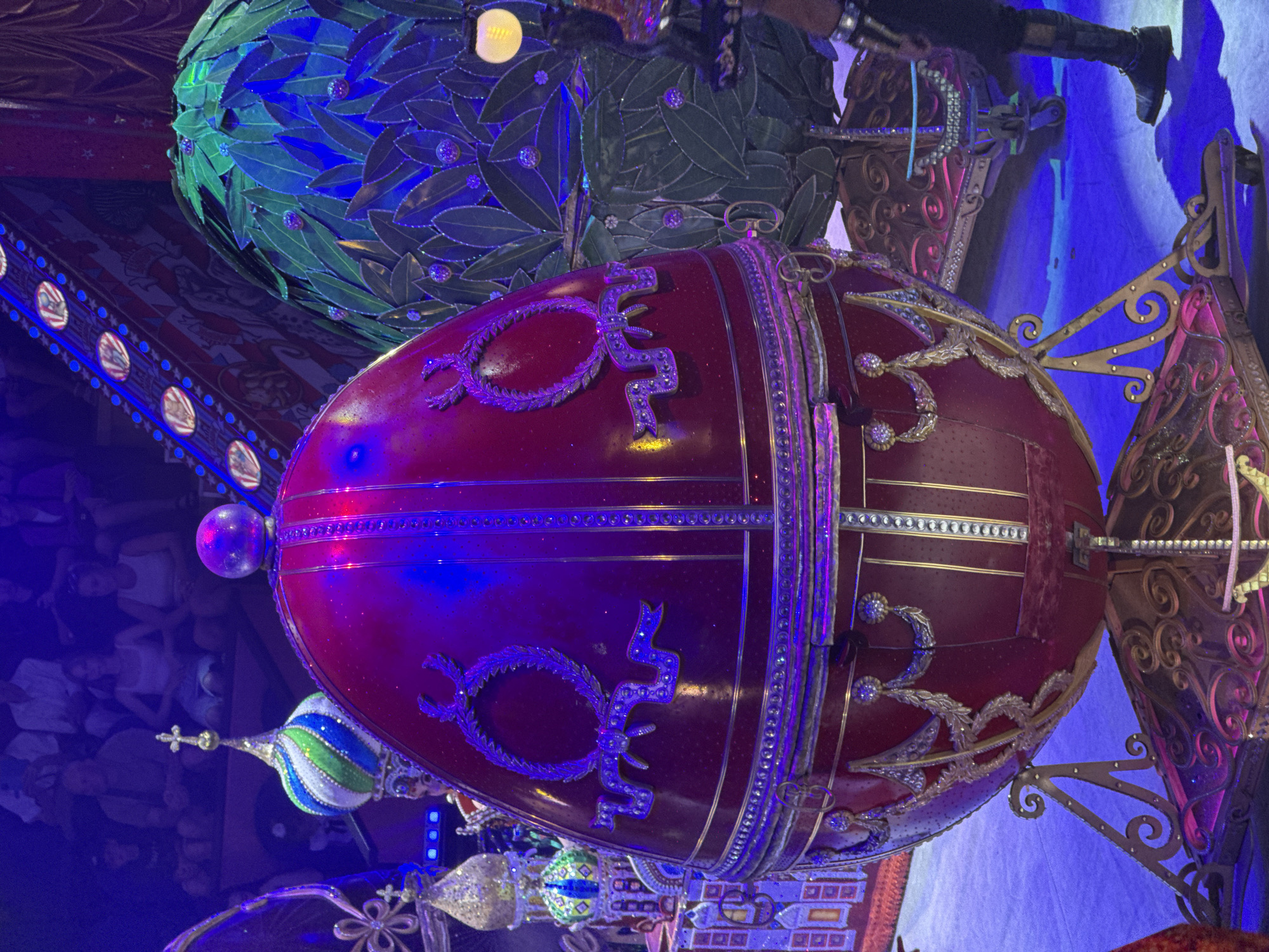 The image depicts a theatrical stage setting, possibly during a performance. The foreground is dominated by a large, ornate, ruby-red egg-shaped object, which appears to be a prop. This object is supported by an elaborate golden stand with swirling metalwork. The egg is decorated with gold trim and intricate patterns, including two oval embellishments that resemble wreaths with ribbons.
Behind the red egg, to its right, is another large prop shaped like a spherical bush or tree covered in dark green leaves, also adorned with small, sparkly purple spheres. The lighting on the stage is predominantly blue and purple, with some yellow accents from a distant light source. In the background, there are glimpses of audience members seated in tiered seating, illuminated by the stage lights. To the left of the red egg, a colorful, domed structure with a cross on top is visible, resembling a miniature Orthodox church, also highly decorated with glitter and jewels. The overall impression is one of fantasy and spectacle.
There is no information in the image that allows for the determination of a specific location in the city of Sochi, Russia.