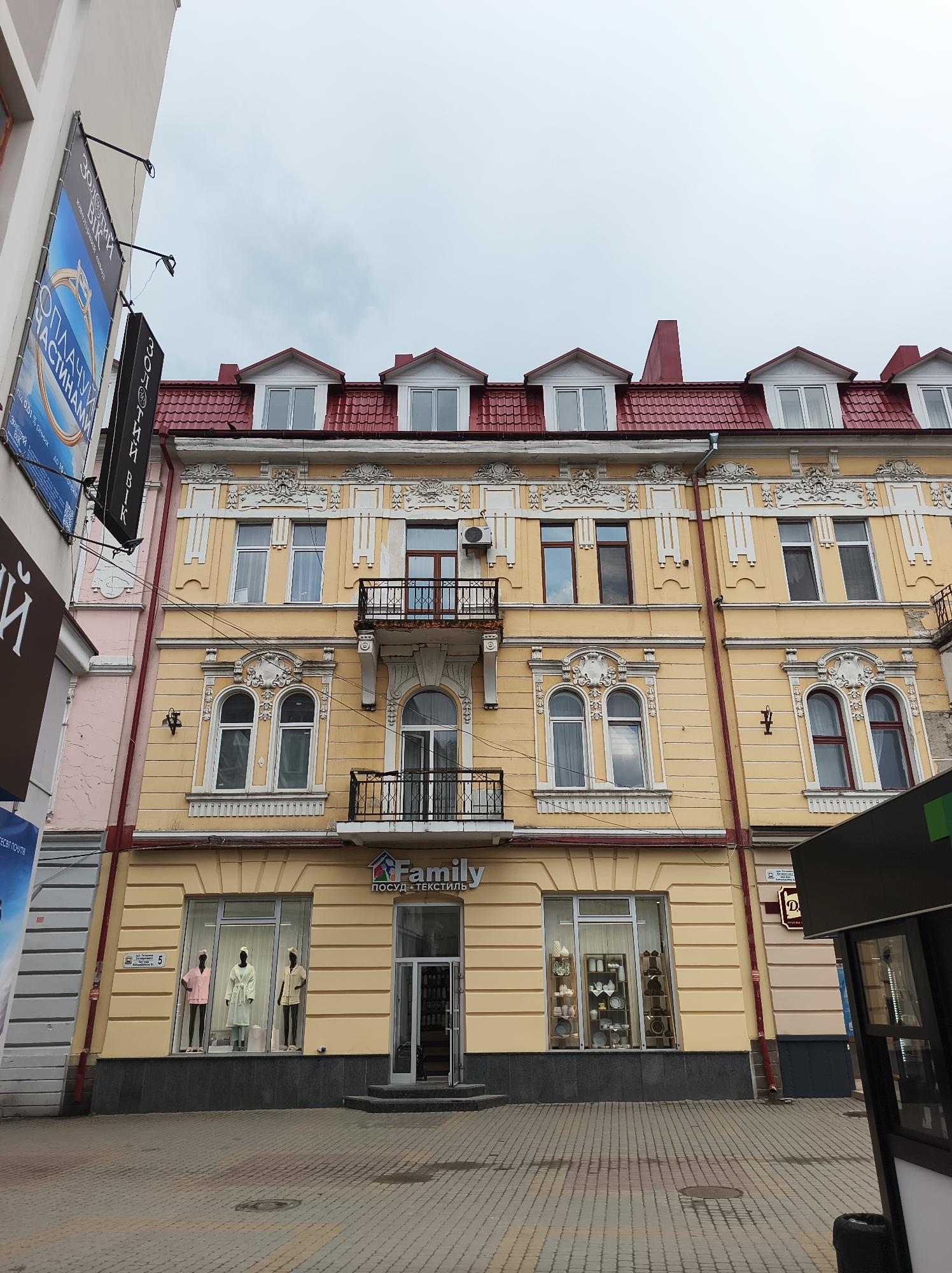 The image captures the façade of a three-story building in Ternopil, Ukraine. The building is painted a light yellow, with white window frames and decorative trim. It has a red tile roof with four dormer windows. On the ground floor is a store named "Family посуд текстиль," which translates to "Family Dishes Textiles" and features display windows with mannequins and various houseware items. There are two balconies on the second floor. A black sign for a jewelry store is visible on the left side of the frame. The street appears to be paved with bricks. The sky is overcast. A bus stop and sidewalk are on the right side of the image.