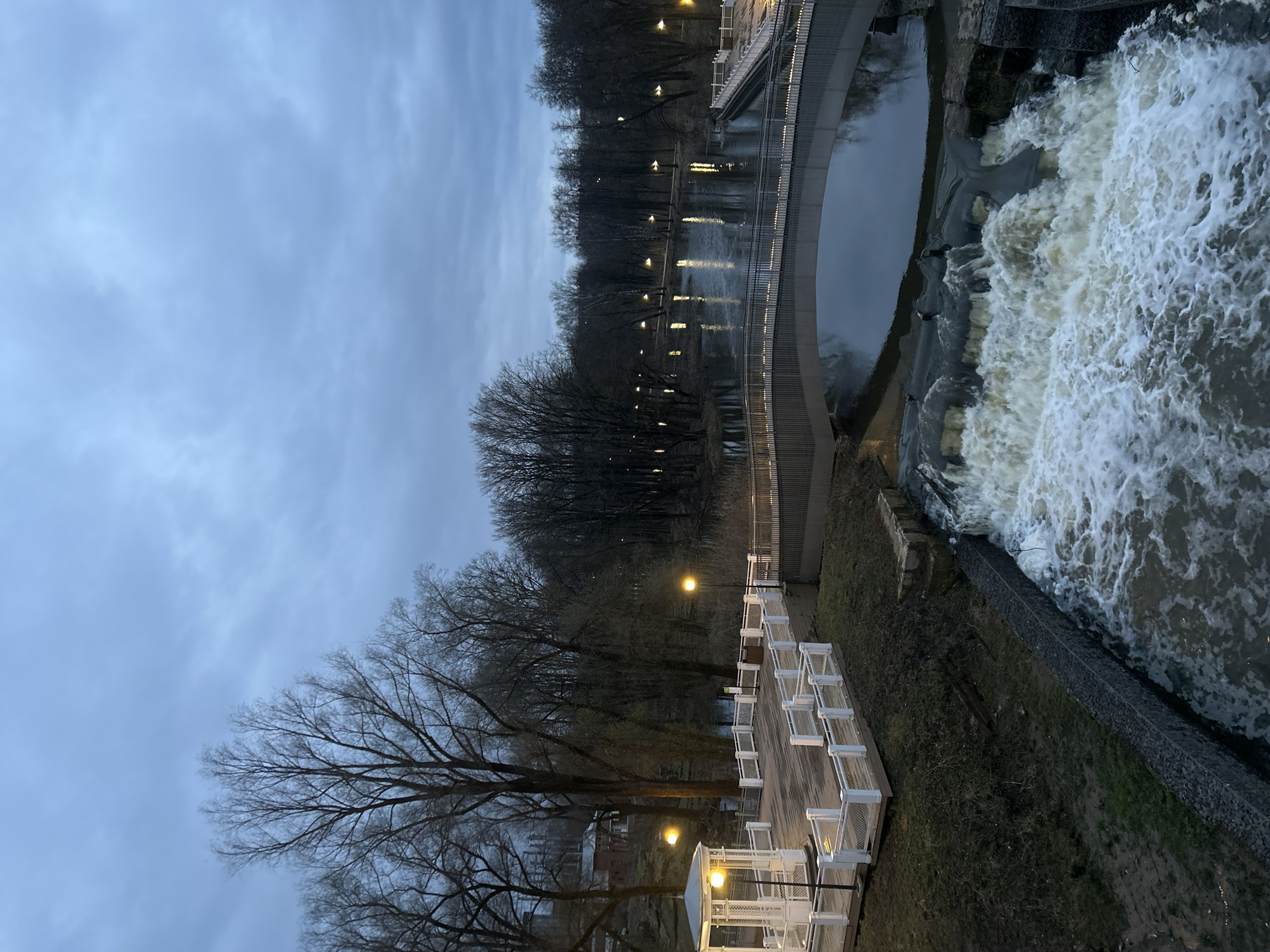The image depicts a nighttime view of a waterfall in Klin, Russia. The water cascades over a constructed dam or weir. A curved pedestrian bridge spans the river above the waterfall. Along the riverbank is a lighted pathway or boardwalk with white railings. Bare trees line the riverbanks, silhouetted against the sky, which shows an overcast, dusk-like condition. Streetlights illuminate the scene. Buildings are visible in the distance.