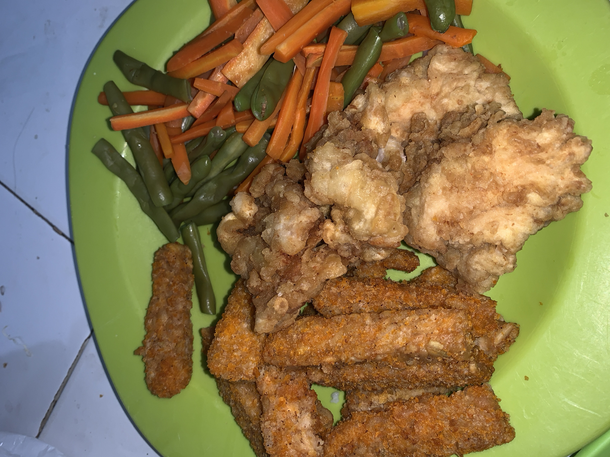 The image displays a plate of food on a surface that appears to be a tiled floor. The plate is bright green and contains a meal consisting of fried chicken pieces, breaded chicken strips, and a side of cooked green beans and sliced carrots. The lighting suggests an indoor setting, possibly a home. There are no people or visible actions depicted in the image, and the environment is limited to the plate and the surface it rests on. The location in Bugih, Indonesia, is inferred from external knowledge of the context in which this image was provided and is not visually determinable from the image itself.