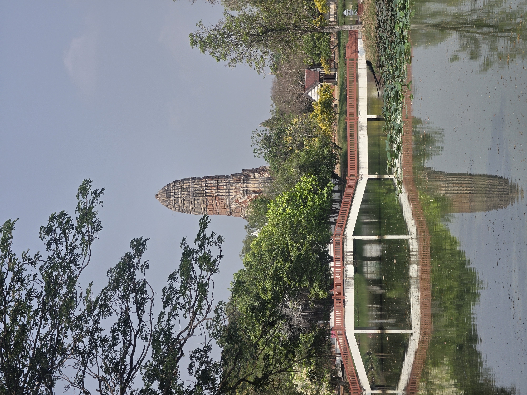 A sideways shot of Wat Phra Ram in the city of Phra Nakhon Si Ayutthaya City Municipality, Thailand, is shown along with its reflection in the water. The temple, which is situated in the upper-center of the image, features a tall, narrow tower-like structure with eroded brick or stone patterns. The tower is surrounded by green trees. At the top-right corner, there is a pond or lake with lily pads floating on the surface and the temple’s reflection. A small building with a white exterior and a dark roof can be seen behind the trees, closer to the upper-right corner. A red and white pedestrian bridge spans over the pond, connecting the foreground and the background. The lower-left corner shows a tree with bare branches and sparse leaves. The sky is a clear light blue, and the overall scene gives a serene and picturesque view of the historic site.