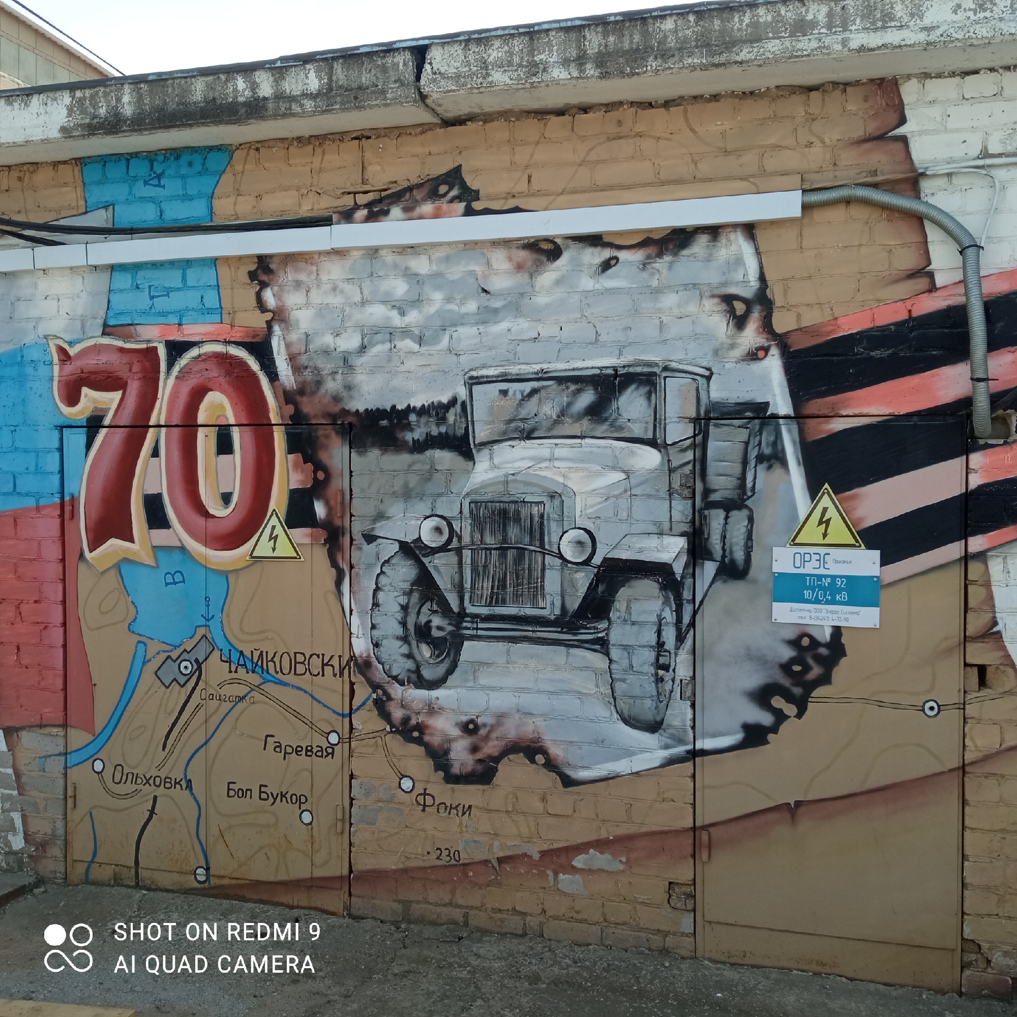 The image depicts a brick wall in the city of Chaikovsky, Russia, adorned with graffiti. The graffiti includes a large red "70", a map featuring the locations of "Chaikovsky", "Saigatka", "Garevaia", "Olkhovka", "Bol Bukor", and "Foki", and a grayscale depiction of a vintage truck. A St. George ribbon is also present. A small metal door with an electrical hazard sign and the text "ОРЭС Прикамья ТП-№ 92 10/0,4 кВ" is affixed to the wall. The text "230" is also present. The concrete pavement is visible at the bottom of the frame. A silver electrical conduit runs along the top of the door.