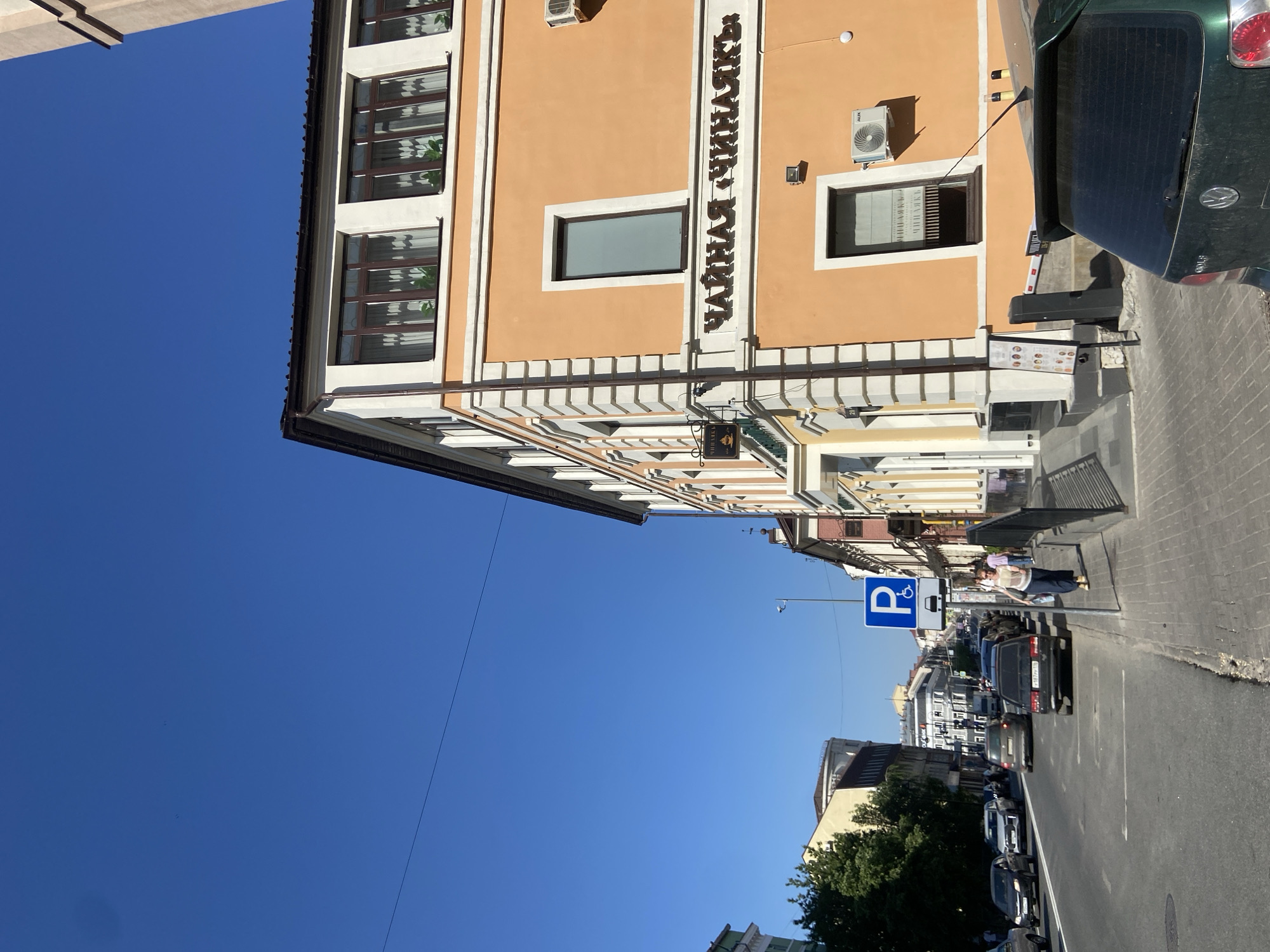 The image shows a street scene in Kazan, Russia, featuring a building with a tan facade and a sign in Cyrillic that reads "ЧАЙНАЯ ЧИЧАЛКА." The building has white trim, multiple windows, and an air conditioning unit mounted on the wall. A green car is parked very close to the building's right corner. On the sidewalk, there's a bench, a menu board, and a person standing near a parking sign marked with a wheelchair symbol (disability parking). The street is lined with parked cars, and additional buildings and trees can be seen in the distance. The sky is clear and blue.