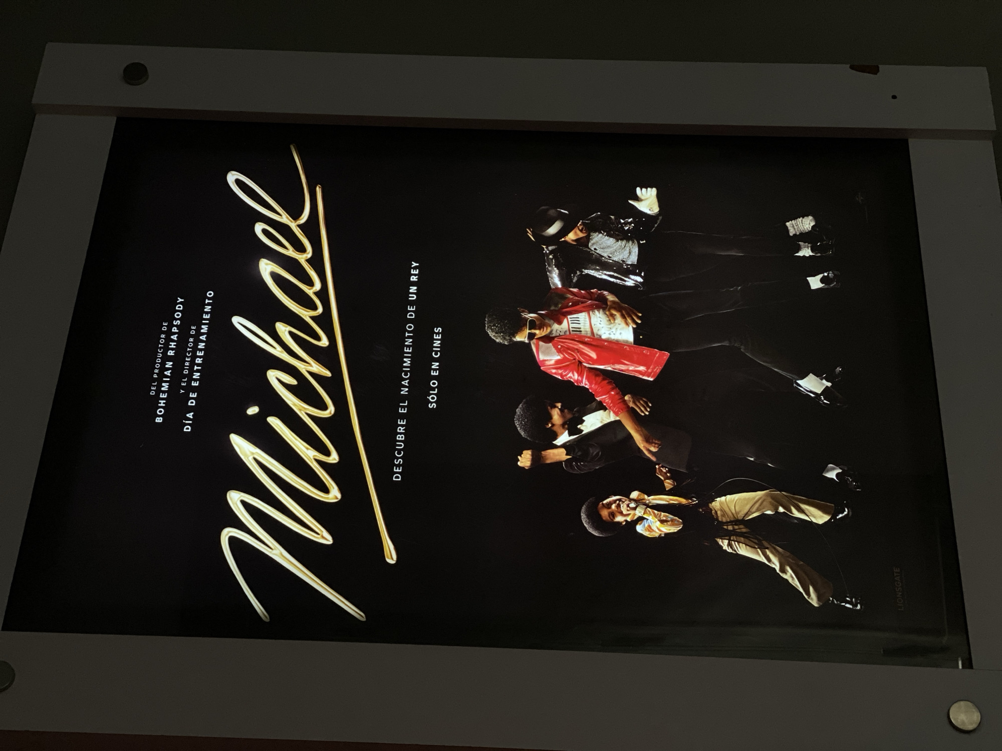 The image shows a framed poster for the movie "Michael," which is about Michael Jackson. The poster features a golden script spelling out "Michael" along the left side. To the right are images of Michael Jackson in various iconic outfits and poses. Text in Spanish advertises the film as being from the producers of "Bohemian Rhapsody" and the director of "Training Day," with the tagline "Discover the Birth of a King" and "Only in Theaters." The Lionsgate logo is visible in the lower right corner. The poster is encased in a light-colored frame with visible screw-like fasteners.