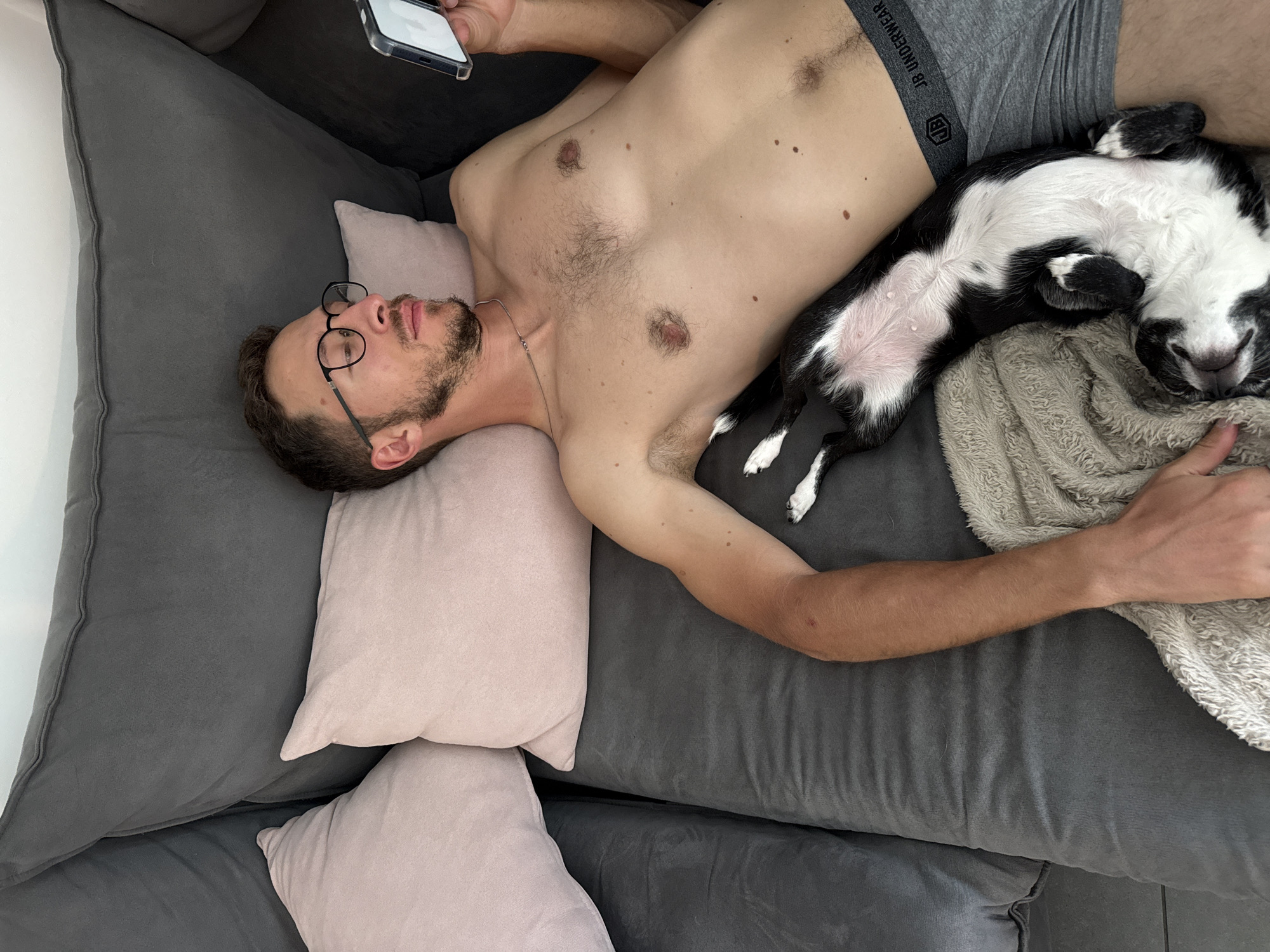 A man is lying on a grey sofa. He is wearing glasses and grey underwear. His chest is bare. He is holding a cell phone in his hand. A black and white dog is lying next to him. The scene appears to be indoors and is not identifiably in Tel-Aviv, Israel.