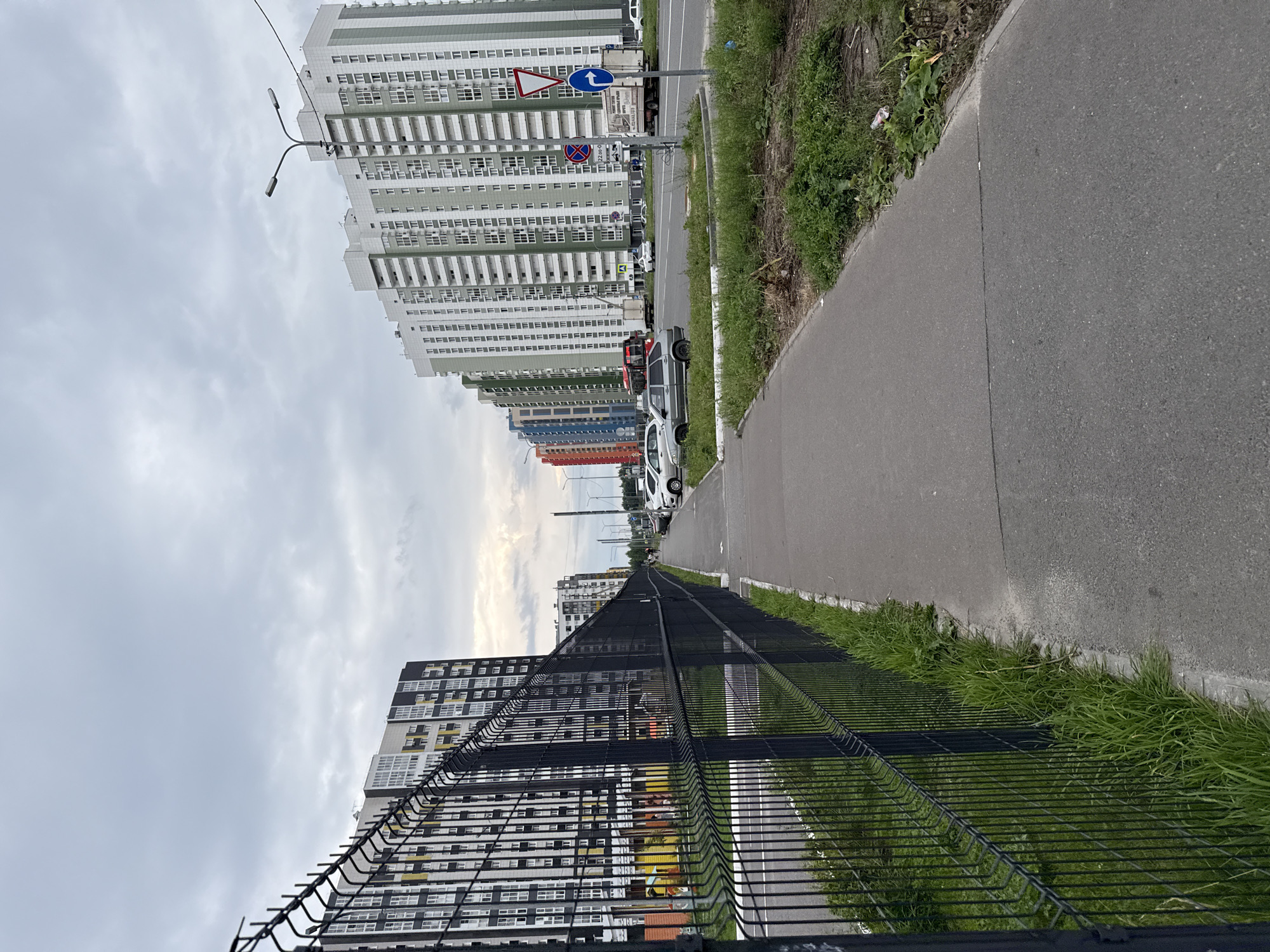 A residential street scene in Kazan, Russia, on an overcast day. A black chain-link fence borders a paved sidewalk on the left, separating it from a grassy area. Beyond the fence, a multi-story building with black, white, and yellow-accented facade rises. Across the street, a row of tall, modern apartment buildings with white and light green facades stretches into the distance. Cars are parked along the street, and a few individuals are visible walking. Traffic signs, including a yield sign and a turn arrow sign, are present. The overall environment suggests a newly developed urban area.