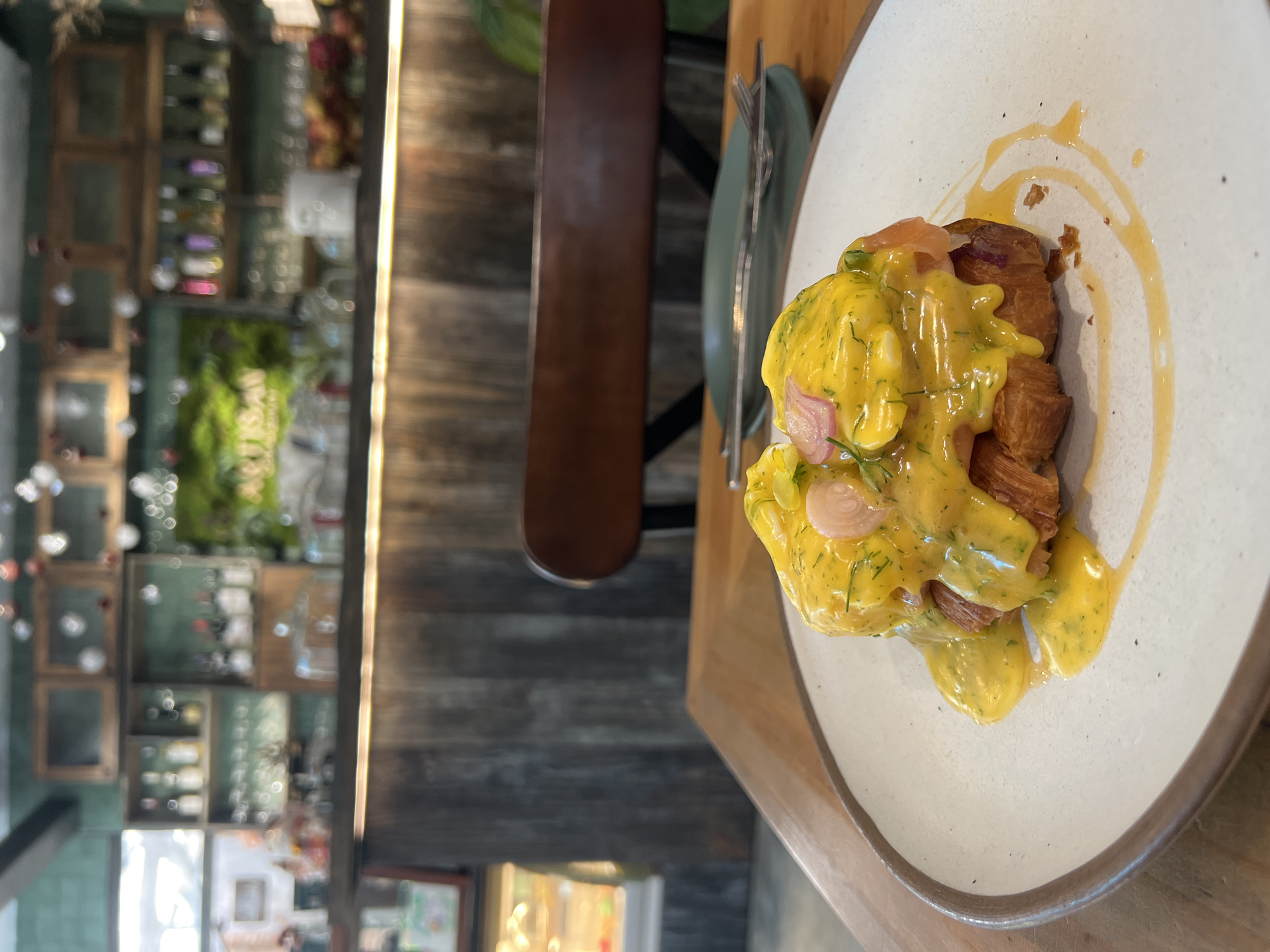 A close-up, slightly rotated shot reveals a plate of food on a wooden table, likely taken in Nha Trang, Vietnam. The dish, prominently displayed on a white speckled plate with a brown rim, features a croissant topped with a yellow sauce, likely hollandaise, garnished with herbs and what appears to be pickled onions and salmon. Utensils are placed neatly to the side of the plate. In the background, a blurred interior scene is visible, showcasing a decorative wall with shelves displaying various items, suggesting a restaurant or cafe setting. The overall scene is well-lit with natural light, creating a vibrant and appealing food photograph.