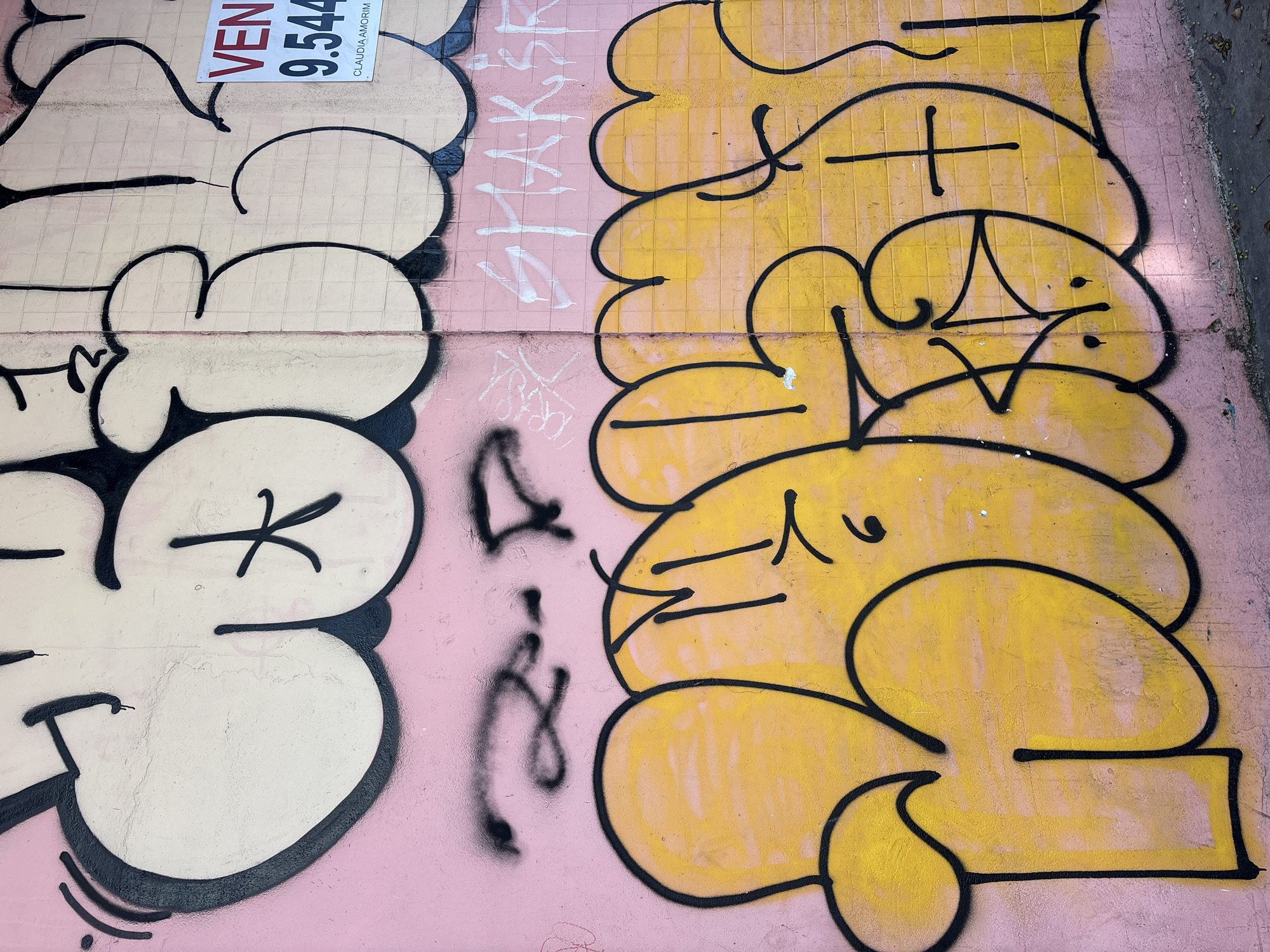 The image depicts a section of an exterior wall in São Paulo, Brazil, covered in graffiti. The wall is painted pink, with tiled sections visible towards the top. Large, bubble-style graffiti tags in white with black outlines dominate the upper portion of the wall, while vibrant yellow graffiti with black outlines fills the lower half. A "VENDA" (for sale) sign with the number "9.544" and the name "CLAUDIA AMORIM" is affixed to the wall on the right side, above the yellow graffiti. Faint black graffiti tags are visible between the white and yellow sections. The ground in front of the wall is paved and shows scattered leaves and debris. The overall scene suggests an urban environment, likely a street or an alleyway, where street art is prevalent.