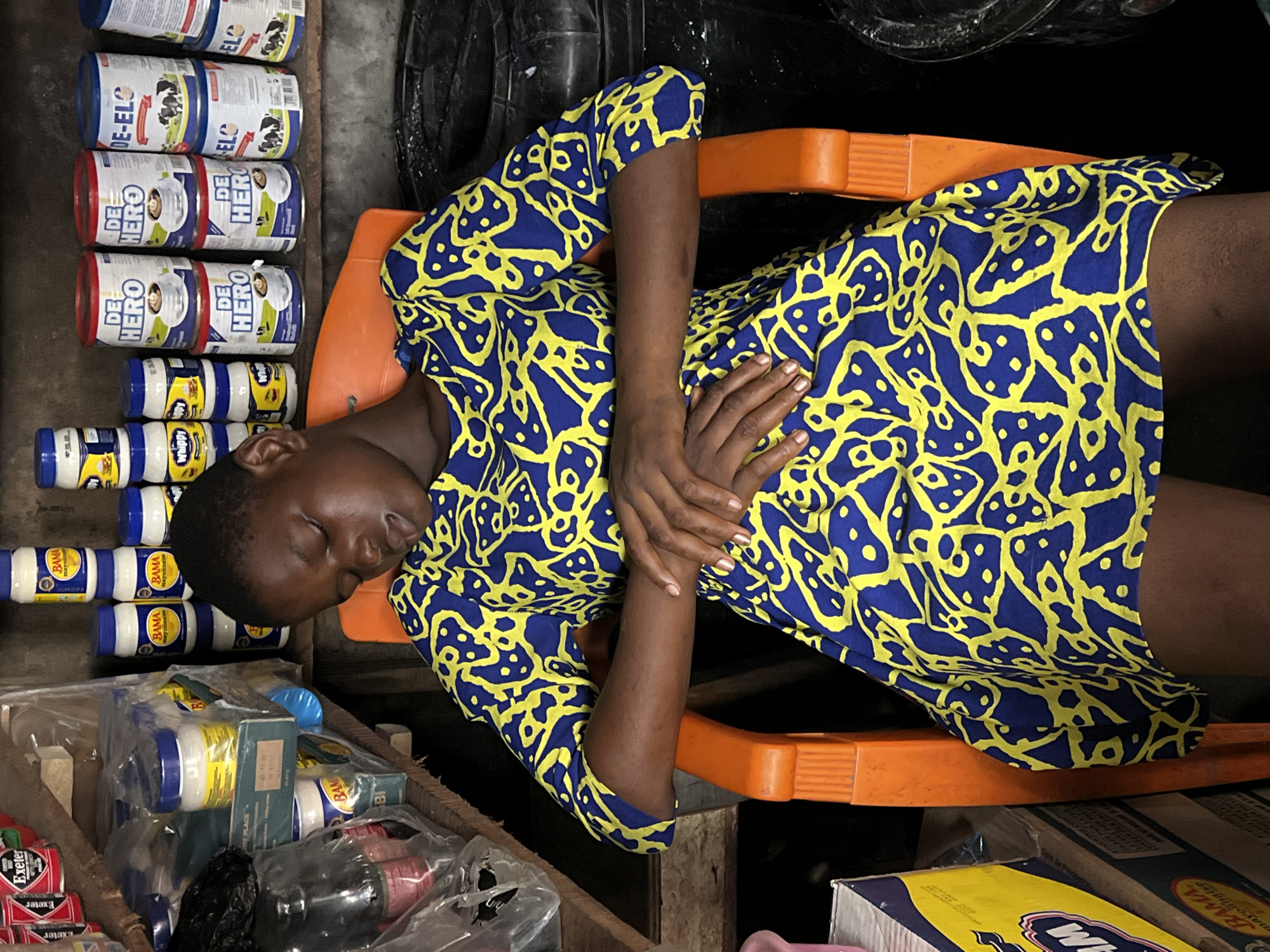 A young woman is sleeping on an orange chair in a shop in an unknown city. She is wearing a blue dress with a yellow pattern. Next to her head are stacks of canned goods: "DE-ELO," "DE HERO," and "Whippy." Below her are boxes of "BAMA" and "Exeter."