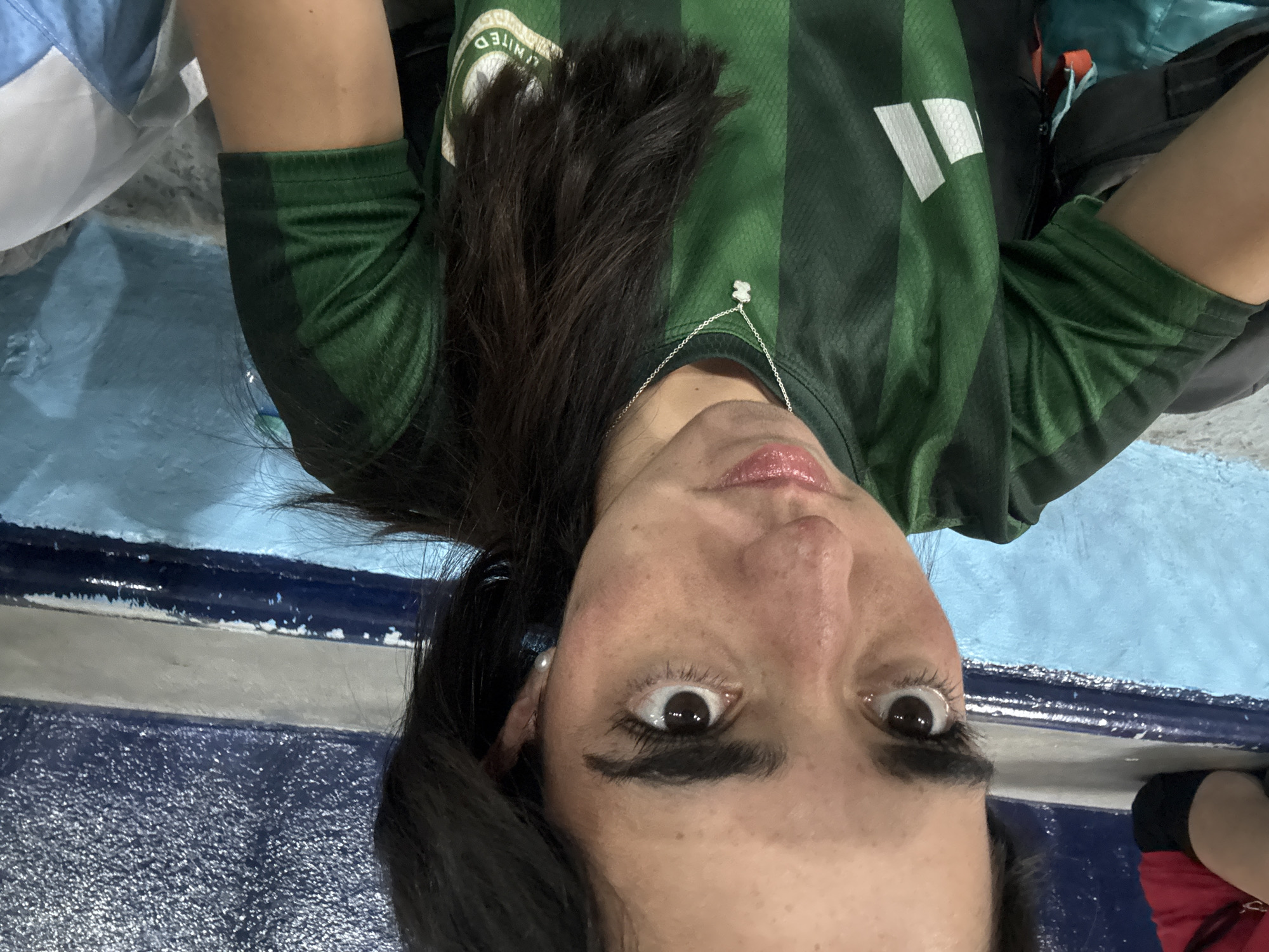 A close-up, low-angle shot features a young woman wearing a green and dark green striped sports jersey, likely at a sporting event. Her dark hair falls over her shoulder, and she looks directly into the camera with wide, dark eyes, conveying a sense of surprise, excitement, or intense focus. Her lips are slightly parted, revealing a glossy pink finish.
She accessorizes with a silver necklace featuring a small, white, bear-shaped pendant and pearl-like earrings. Her jersey displays a white logo with two vertical bars on the right shoulder/chest (viewer's left) and a partially visible circular logo that reads "UNITED" on the left chest (viewer's right).
The background shows a dark blue, textured structure, possibly stadium seating or a wall, with a lighter blue surface beyond it, suggesting an indoor or stadium environment. A light blue and white garment is partially visible on the left side of the frame. The image likely captures a personal moment or selfie at a sporting event, with the context being Mexico City, Mexico.
