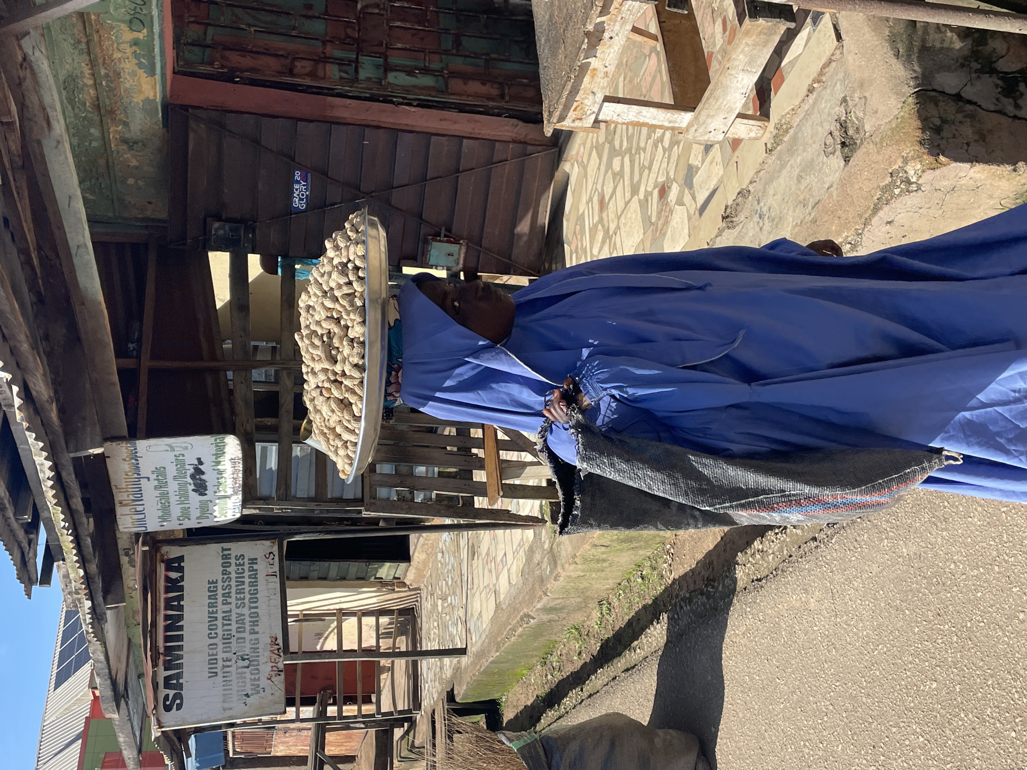 In Jos, Nigeria, a woman dressed in a blue hijab and robe balances a large metal tray of peanuts on her head. She walks on a paved path alongside a concrete drainage ditch, carrying a large woven bag over her shoulder. The background features a row of shops with corrugated metal roofs, some displaying signs in English. One sign reads "SAMINAKA VIDEO COVERAGE 1 MINUTE DIGITAL PASSPORT NIGHT AND DAY SERVICES WEDDING PHOTOGRAPH". Another sign advertises "Uncle Mathu Shoe Special Wholesale/Retails, Shoe Making/Repairs, Dyeing, Sewing, Sales of Material". Solar panels are visible on one of the rooftops. The ground is a mixture of paved areas and dirt. Sunlight casts shadows across the scene, indicating a daytime setting.