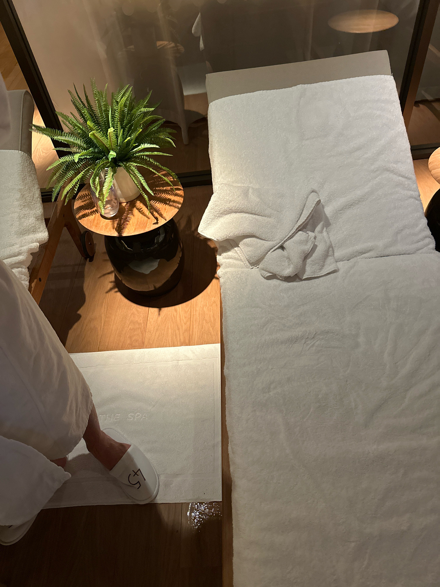 In a spa room in London, United Kingdom, a person wearing a white bathrobe and white slippers with "45" printed on them is stepping onto a white mat with "THE SPA" embossed on it. The mat is placed on a wooden floor. To the left of the mat, a small round wooden table holds a potted green fern in a white vase, with a reflection of soft lighting on the surrounding surfaces. To the right of the table, a white massage bed is covered with clean white towels, and a folded towel rests on top. The room appears to be dimly lit with warm lighting, creating a tranquil atmosphere.