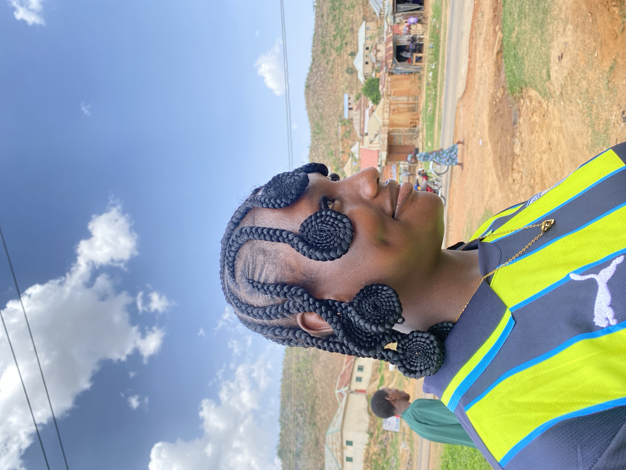 A woman with braided hair adorned with coiled extensions stands outside on a partly sunny day in Gwong, Nigeria. She wears a black, neon green and blue striped sports shirt and a gold necklace. The background shows buildings and a hillside, suggesting a residential area, and a person in a green shirt is partially visible behind her. A road is present on the right side of the image.