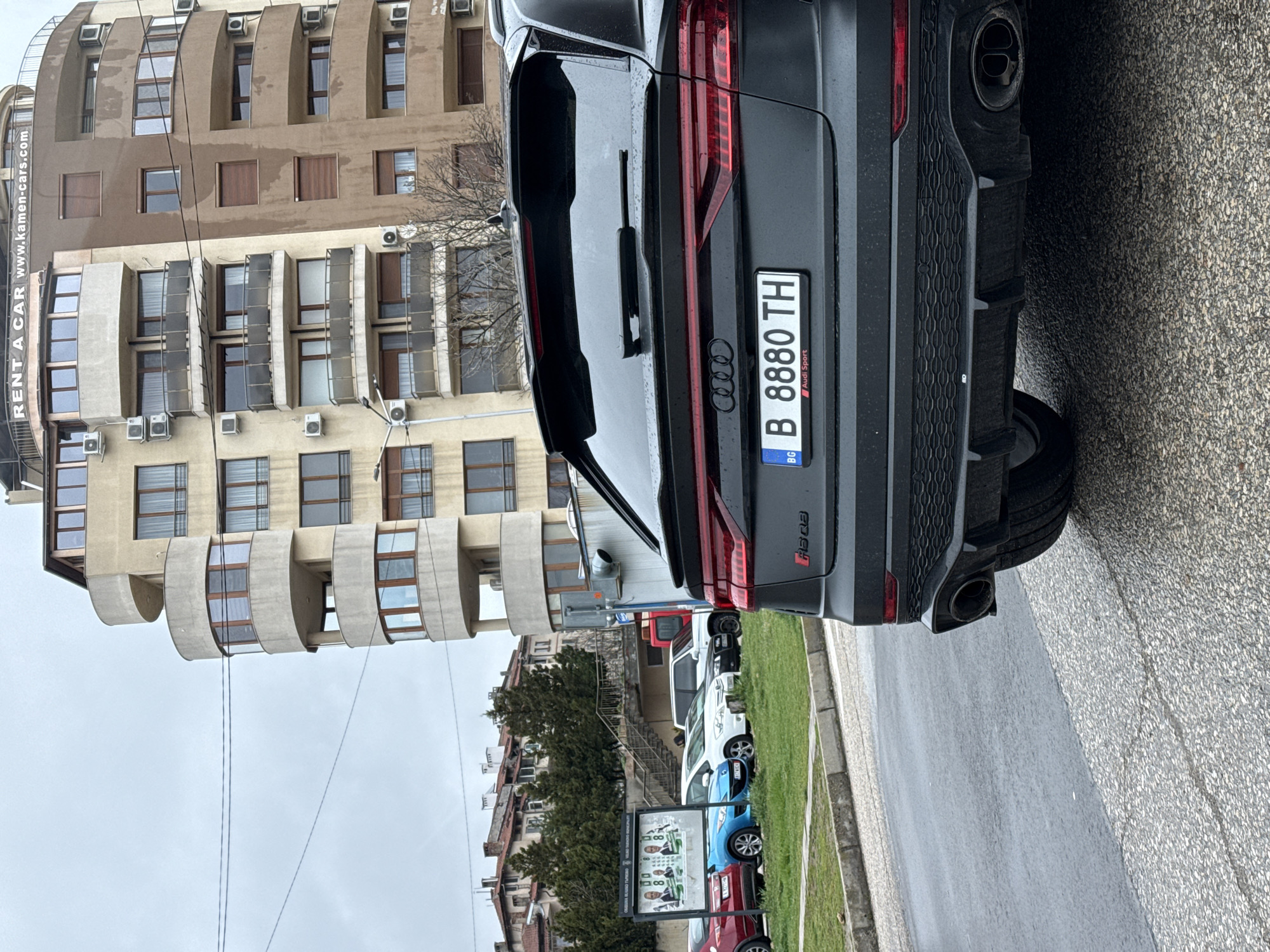 A dark gray Audi RSQ8 SUV is parked on a paved street in Veliko Tarnovo, Bulgaria. The license plate number is B 8880 TH. In the background is a multi-story apartment building with a "RENT A CAR" sign. Other parked vehicles are visible, along with greenery and a political campaign sign on a stand. The weather appears overcast.