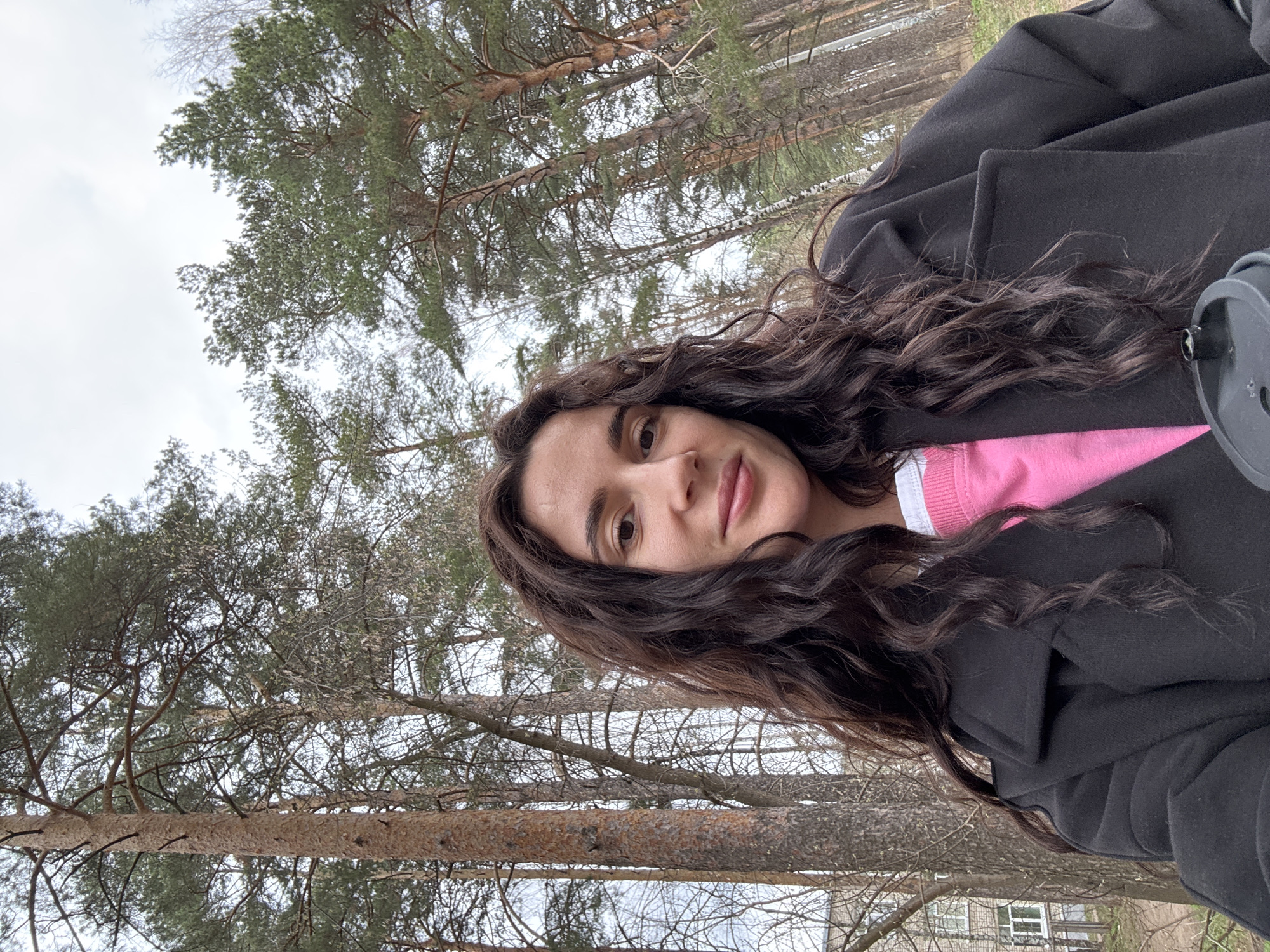 A woman with long, dark, wavy hair is the primary subject in this outdoor selfie. She is wearing a black coat over a pink top with a white collar, and part of a grey cup is visible near her right shoulder. The background shows a forest or wooded area with tall pine trees, their branches stretching towards a cloudy sky. In the lower-right corner, a portion of a building with a window is visible, suggesting the location may be near an urban area. Based on visual cues, the location is consistent with a park or green space within a temperate climate region. The exact location in Chaikovsky, Russia cannot be determined from the image alone.