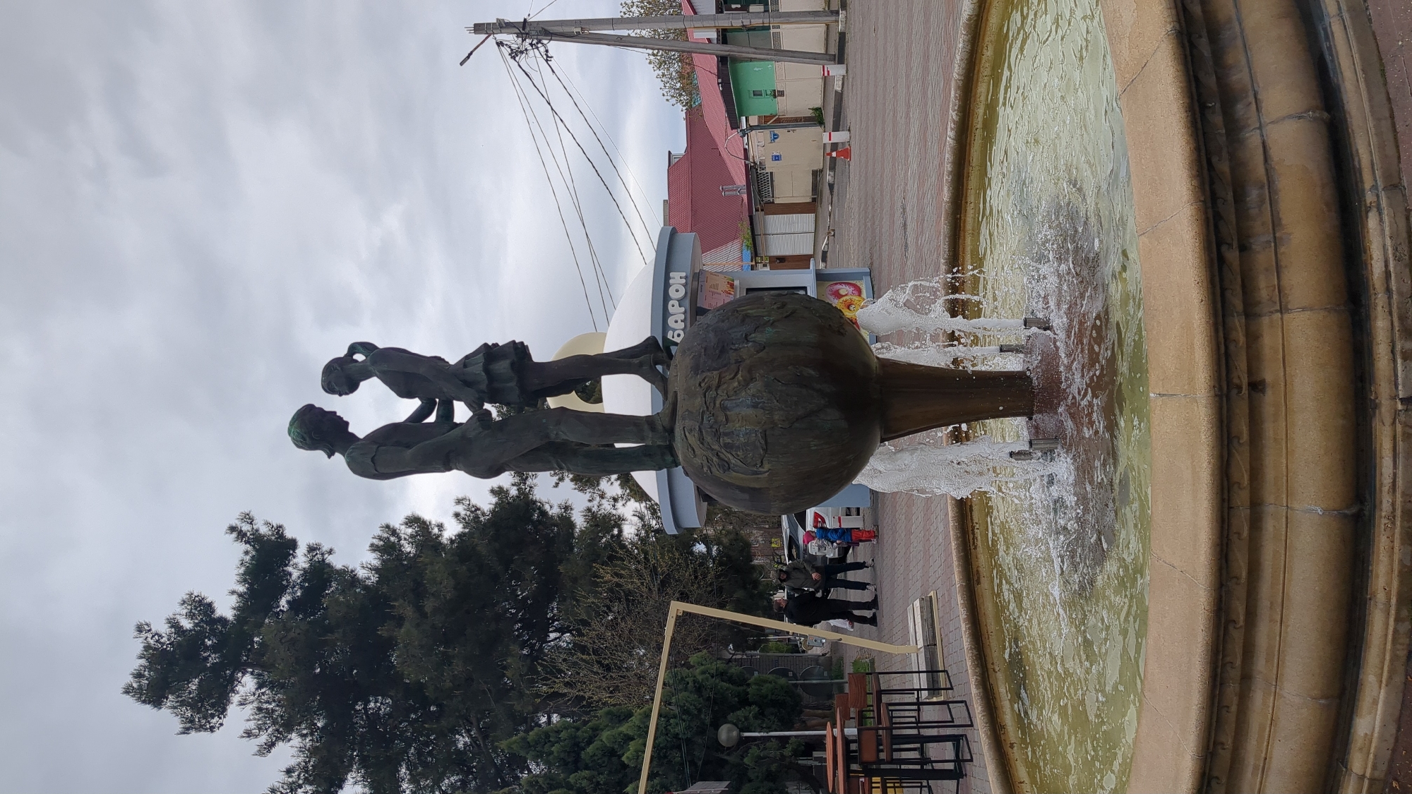 The image shows a sculpture of two figures standing on top of a globe-shaped fountain in Gelendzhik, Russia. The sculpture is made of dark metal. Water is flowing from the fountain into a layered stone basin. Behind the fountain is a building with the word "БАРОН" on its facade. A utility pole stands behind the building. Several people stand on a pedestrian walkway near the fountain. Black metal tables and chairs are stacked near the bottom of the frame. A tree with dark green leaves is visible on the left. The sky is overcast.