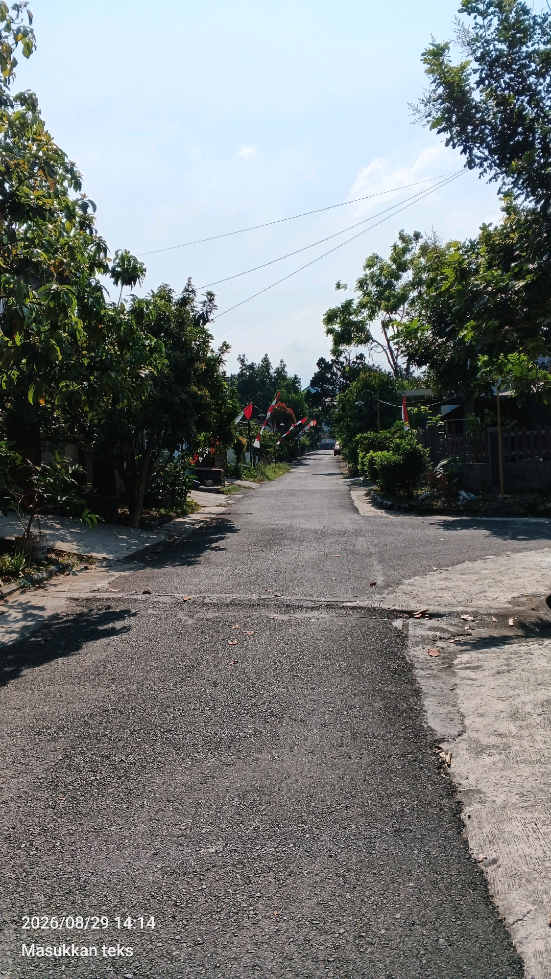 The image depicts a paved road in a semi-rural area, possibly a village or a suburban street, in Sumberporong, Indonesia. The road appears to be asphalt, with some concrete sidewalks or verges. Lush green trees line both sides of the road, casting shadows on the pavement. Indonesian flags, red and white, are visible strung along the road on one side, suggesting a local celebration or national holiday. The sky is bright blue with a few scattered clouds. The scene is quiet and serene, with no people or vehicles immediately visible. The overall impression is one of a typical day in a residential area of Indonesia, bathed in sunlight.