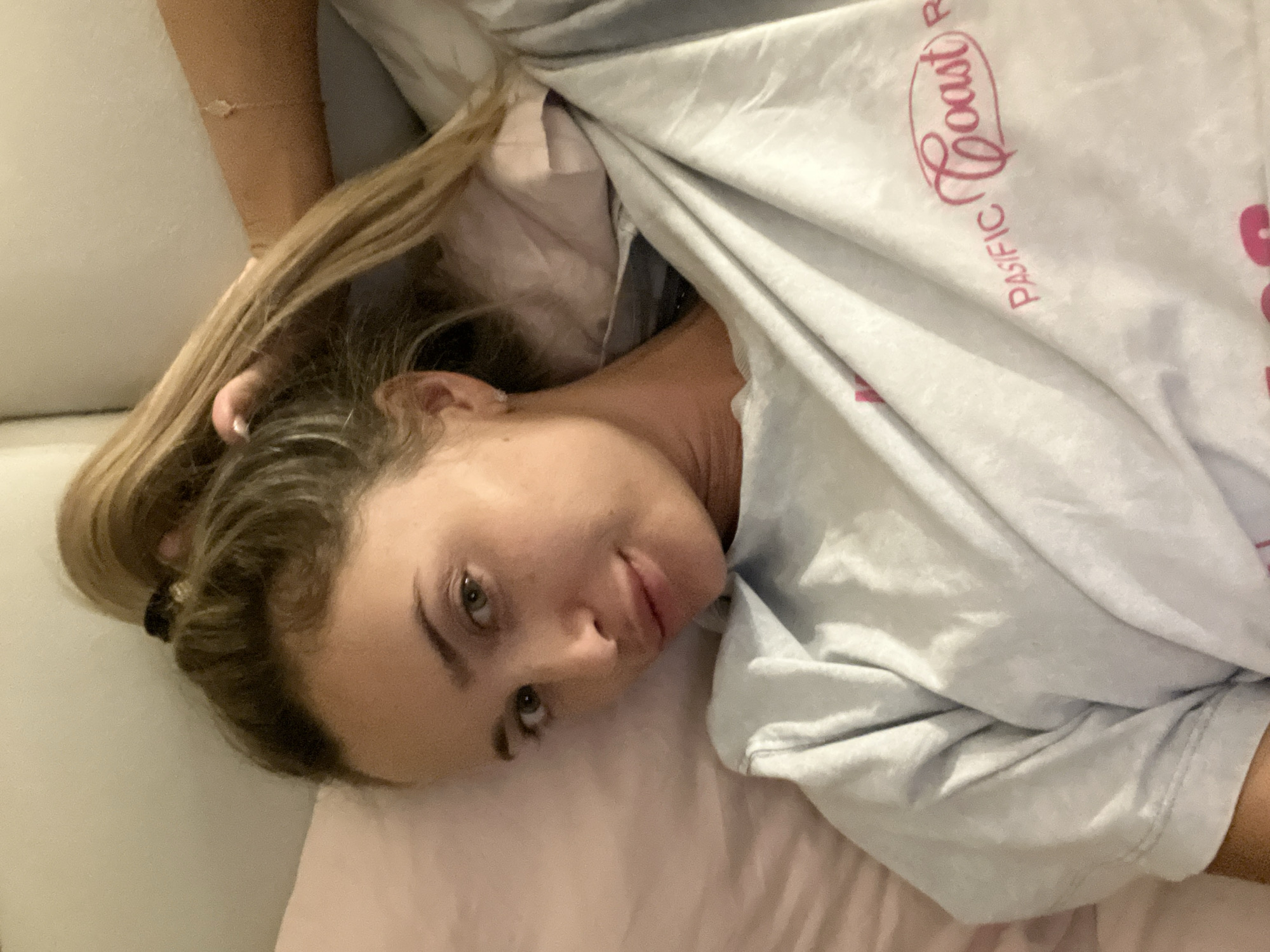 A young woman with blonde hair in a high ponytail lies on her side on a bed, looking towards the camera. She is wearing a light blue t-shirt with pink lettering that reads "Pacific Coast". Her right arm is bent, and her hand is near her head. The background consists of pillows and bedding. The scene is likely indoors, in a bedroom. There is no indication of the specific location within Sharm El Sheikh, Egypt.
