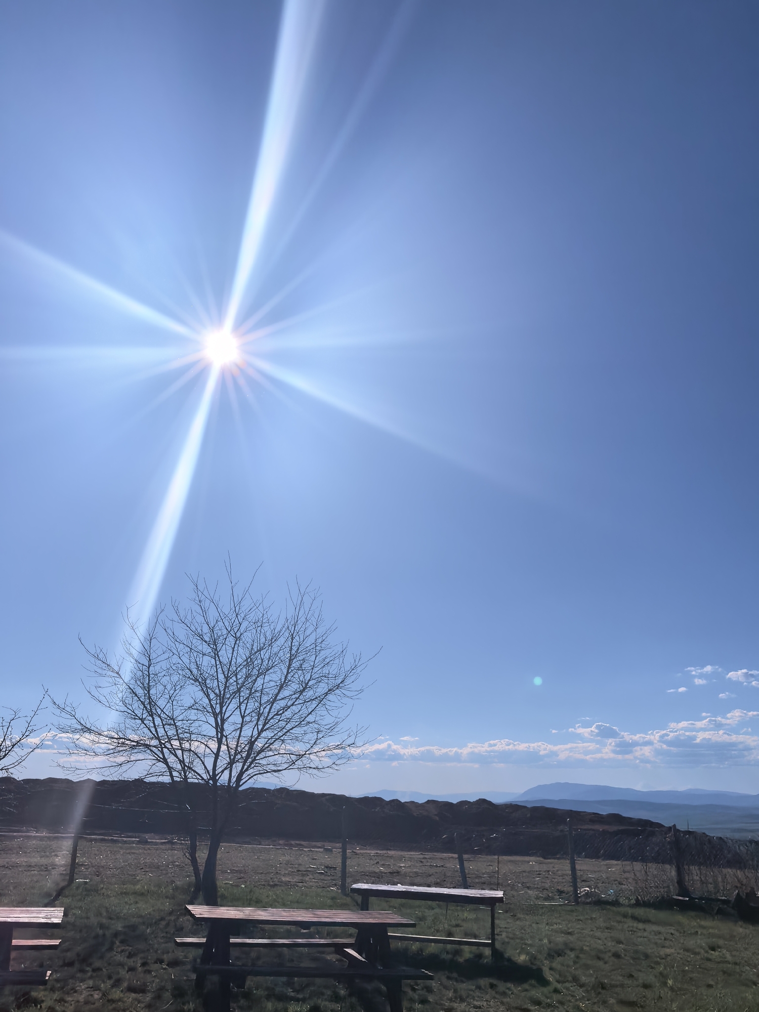 The image captures a rural landscape under a bright, clear blue sky. The sun, positioned high in the upper left quadrant, radiates intense light rays. In the foreground, there are several wooden picnic tables situated on a grassy area. A bare tree stands near the picnic tables. A wire fence runs across the midground, separating the grassy area from a more barren field. Beyond the fence, there's a low ridgeline and distant mountains. Some clouds are visible on the right. The location is Unknown, Unknown.