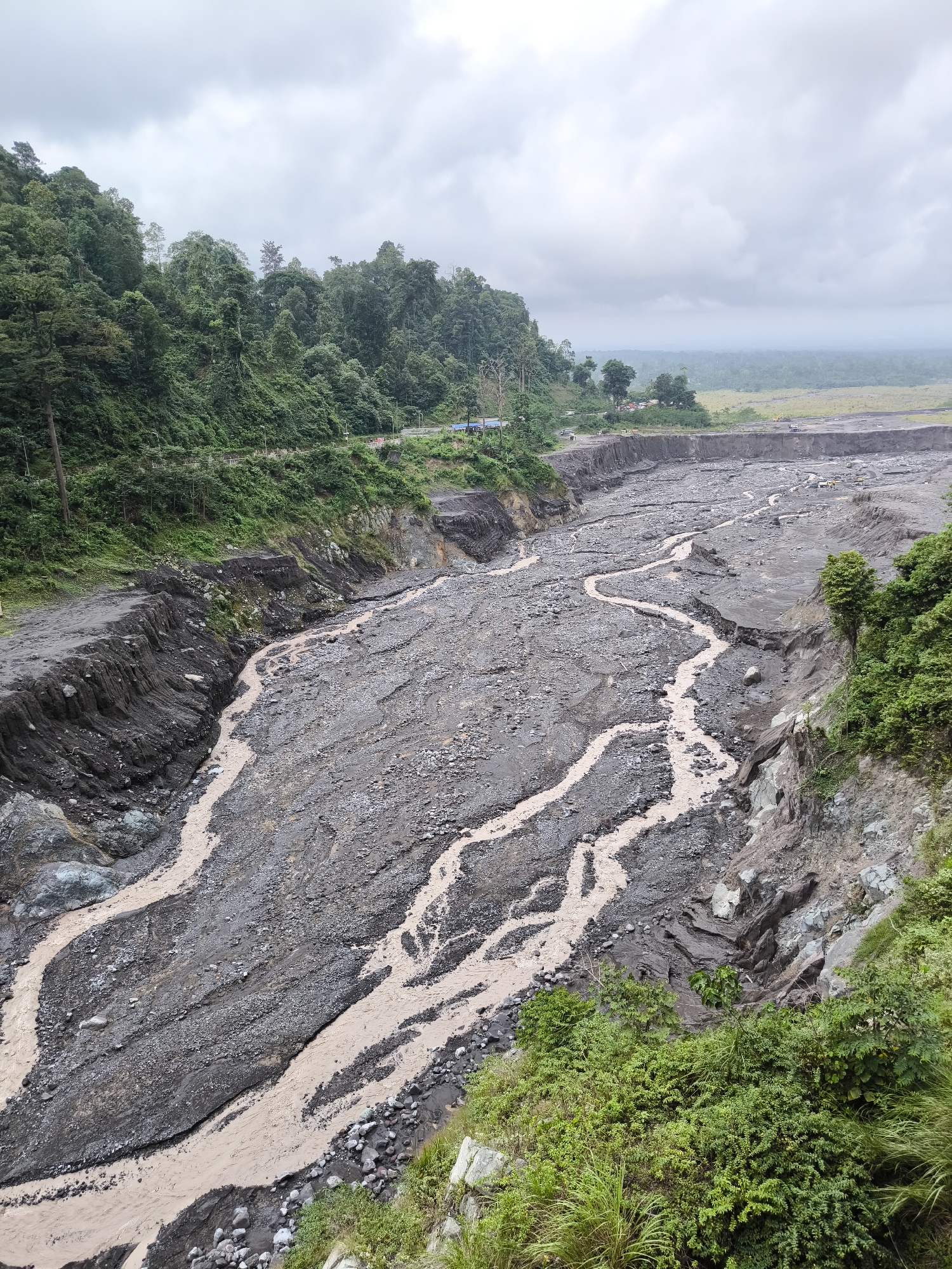 This image shows a wide river valley filled with dark gray volcanic sediment and rocks, carved by a muddy brown river flowing through it. The river has multiple channels that snake across the debris field, indicative of flash floods or lahars. Steep, vegetated hillsides covered in dense green foliage rise on either side of the valley. In the distance, the valley opens up to a hazy landscape with fields and more vegetation under an overcast sky. Evidence of human activity is visible along the top of the left hillside, including a road, streetlights, and what appear to be parked vehicles and some structures. The overall scene depicts a dramatic, geologically active landscape near Supiturang, Indonesia.