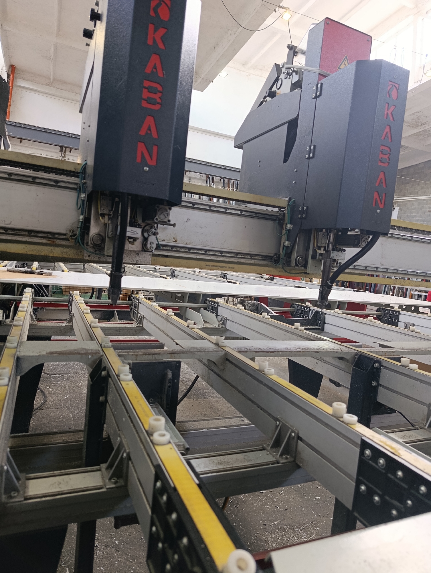 An industrial cutting machine with the brand name "KABAN" is shown from a low angle. The machine is positioned over a conveyor system, and a long, flat white material is laid out on the conveyor. The machine's cutting head is extended and appears to be in a ready or active position. The background consists of a factory or workshop environment, with exposed pipes, conduits, and ceiling structures. The floor is concrete and has some debris scattered on it. The overall scene suggests a manufacturing or fabrication setting.
