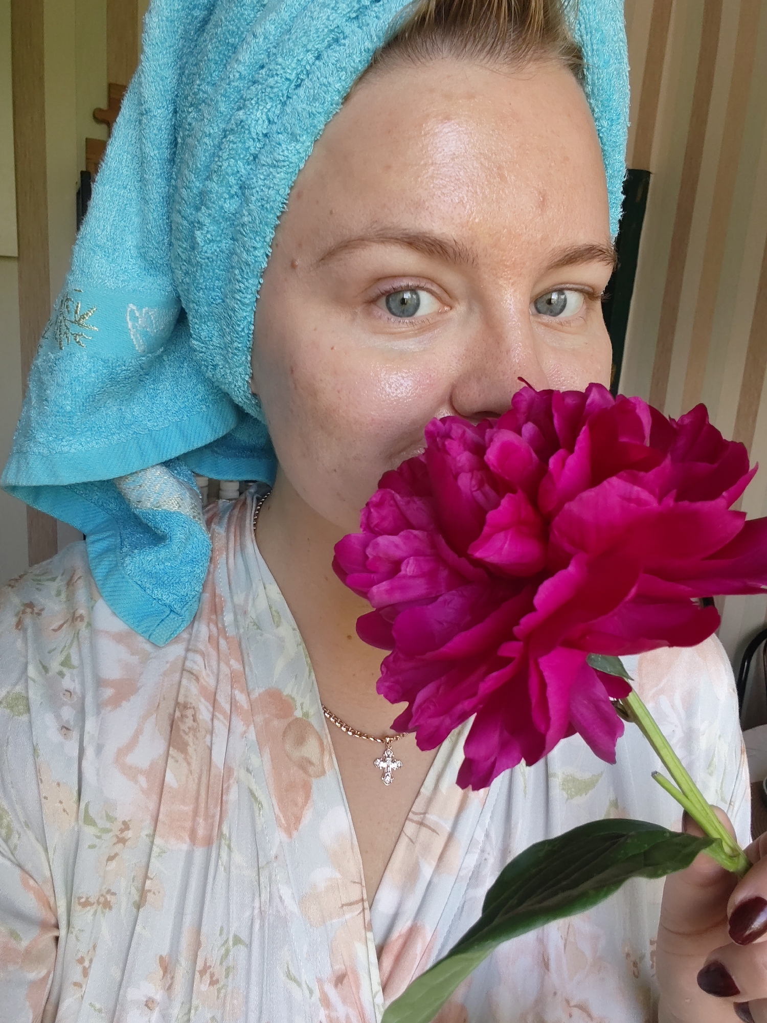 A medium close-up shot shows a woman with blue eyes and light skin, her face partially obscured by a vibrant pink peony flower. She is wearing a light blue towel wrapped around her head, and a robe with a floral print featuring pink and peachy blossoms. A delicate gold chain with a cross pendant rests on her chest. The background consists of striped curtains, suggesting an indoor setting, possibly a residential apartment in Izhevsk, Russia. The lighting appears natural and soft, highlighting the textures of the towel, flower petals, and the woman's skin.