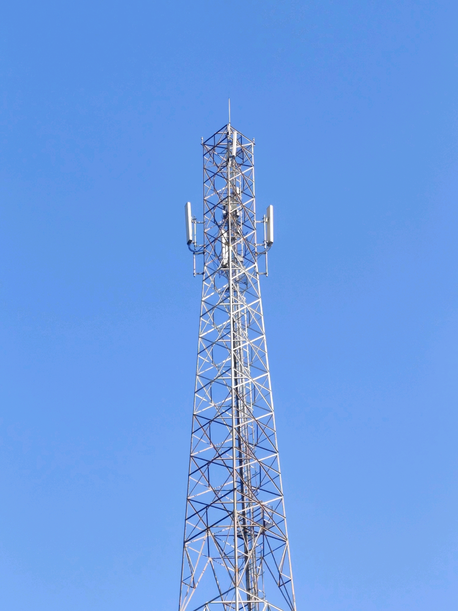 A tall, triangular steel lattice tower stands against a clear blue sky. The tower has several antennas mounted on its sides, with rectangular panels facing outwards. A narrow ladder runs up the length of the tower. The tower is a telecommunications tower, commonly used for mobile phone signals. The image is taken from below, looking up at the tower. The location is the city of Tirtomulyo, Indonesia.