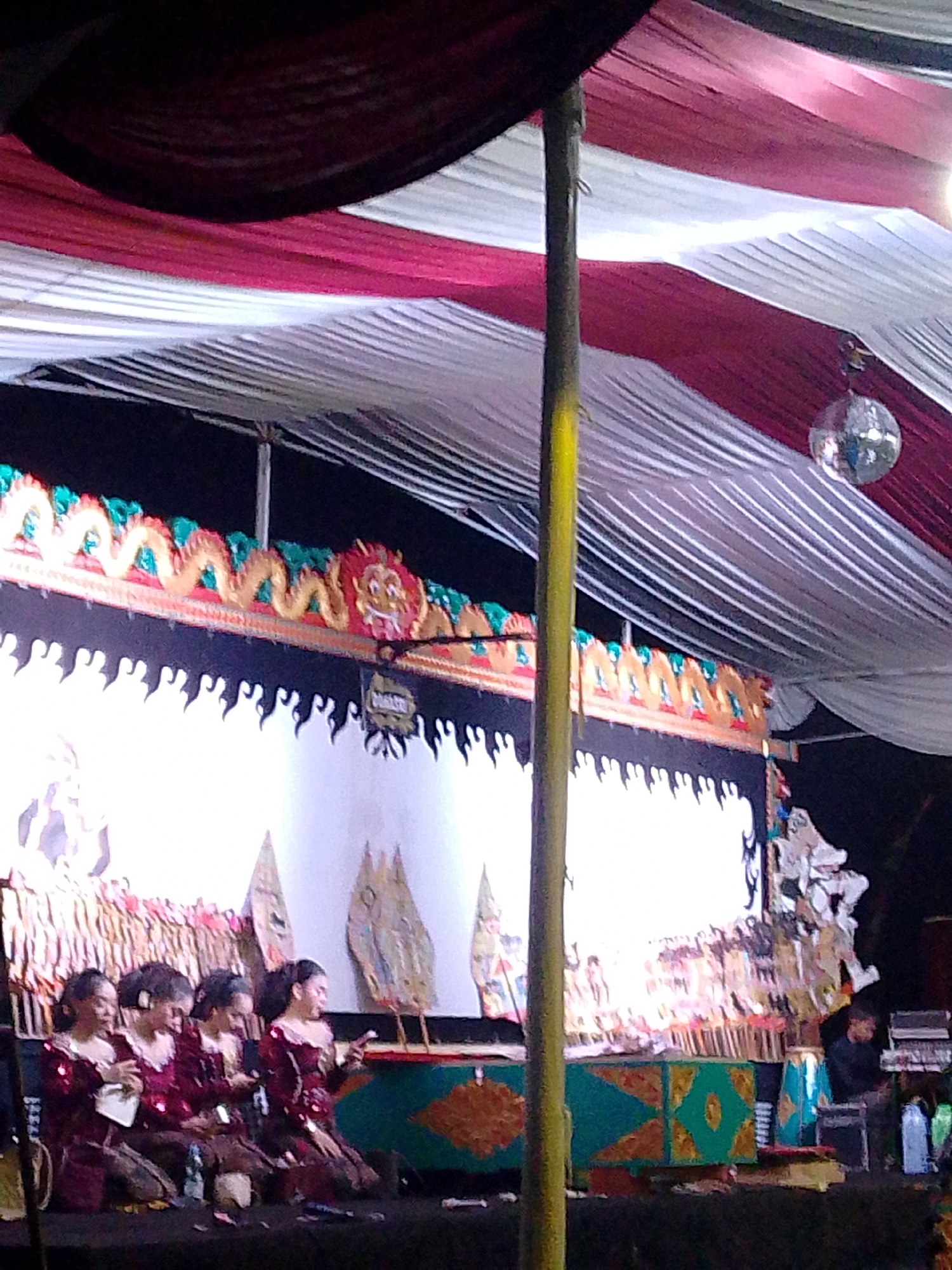 In Wanareja, Indonesia, a group of four young women in ornate, dark red traditional clothing are seated on a dark stage, looking towards a traditional Wayang Kulit shadow puppet performance. The women appear to be performers or audience members attending the event. The stage is set up with a large white screen on which the shadow puppets are projected. The backdrop above the screen is decorated with intricate carvings of dragons and mythical creatures in vibrant colors, including gold, red, and green, suggestive of a cultural performance. A disco ball hangs from the draped ceiling of red and white fabric, adding an unexpected touch to the traditional setting. The overall scene suggests a lively evening cultural event taking place outdoors, possibly a festival or celebration.