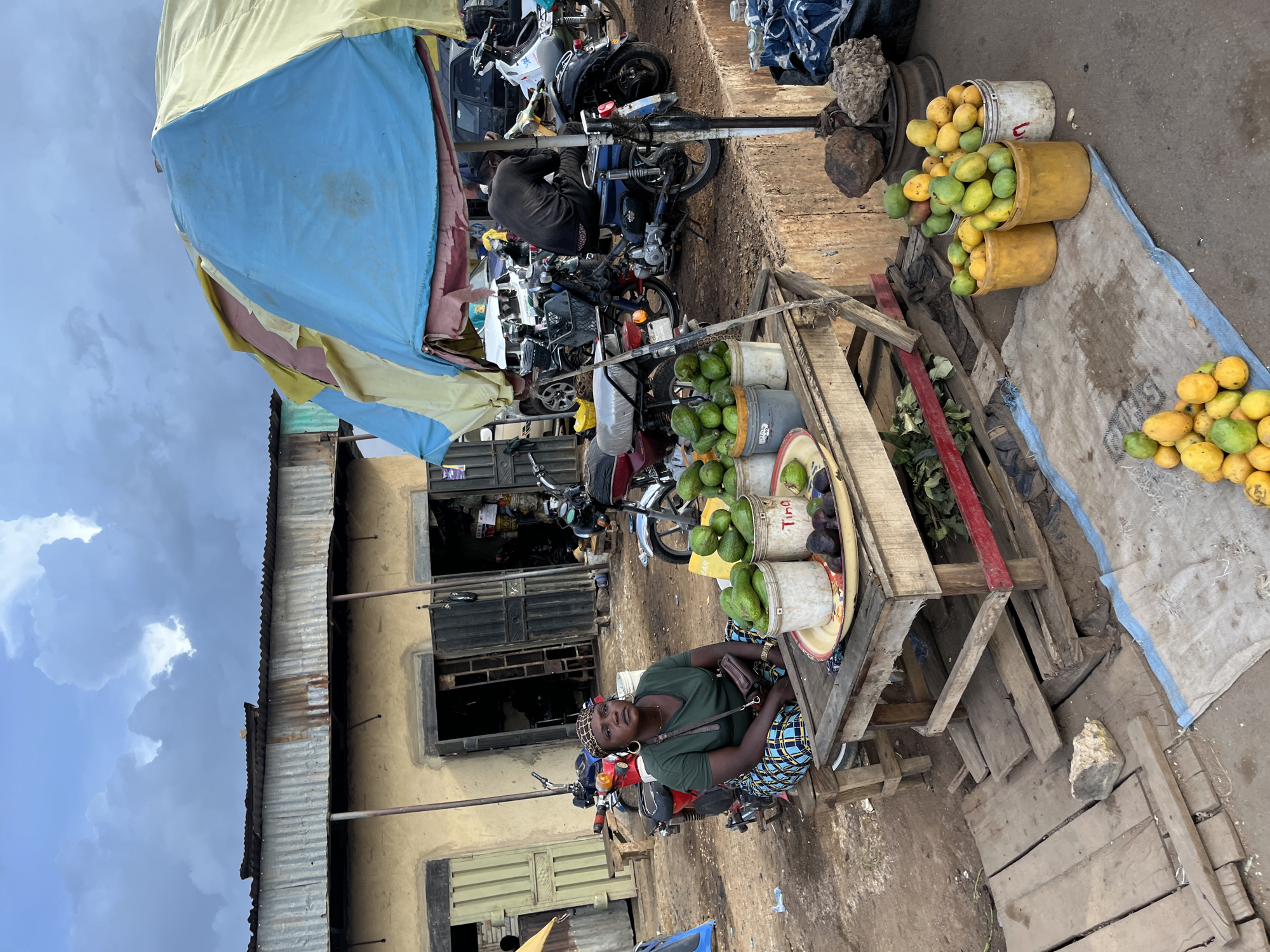 A woman is sitting on a wooden stool in front of a table in an outdoor market in Rafin Par, Nigeria. She is wearing a green t-shirt, a patterned headscarf, and a blue and white checkered skirt. She has a small brown purse slung across her body. In front of her, on a wooden table, are several buckets and a tray filled with avocados and some dark-colored fruits, possibly plums or prunes. To the right of the table, on the ground, are several buckets and a pile of mangoes. In the background, there are several motorcycles parked and a building with corrugated metal roofing and open doorways. A large, colorful umbrella, made of yellow, blue, and pink fabric, is providing shade over part of the market. The ground is dusty earth. The sky is cloudy.