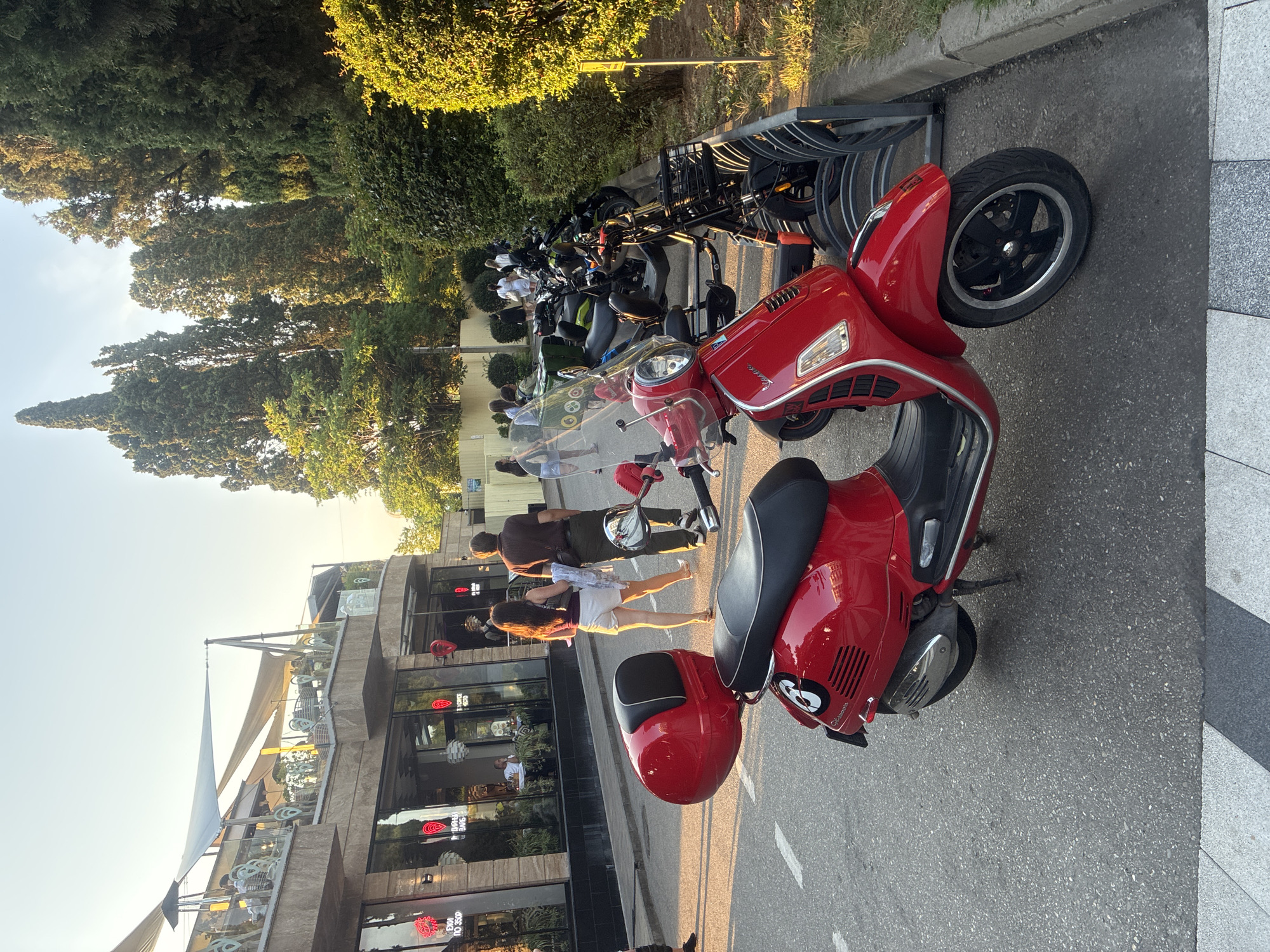 The image depicts a street scene in Sochi, Russia, with a focus on a red Vespa scooter parked on the left side of the frame. In the background, a modern building with a cafe or restaurant is visible, featuring outdoor seating under awnings. Several people are walking on the sidewalk in front of the building. To the right of the scooter, a rack of parked electric scooters is present, with a variety of models visible. Lush green trees and bushes line the right side of the street, suggesting a well-maintained urban environment. The lighting indicates it is late afternoon or early evening, with a warm, golden hue cast by the setting sun.