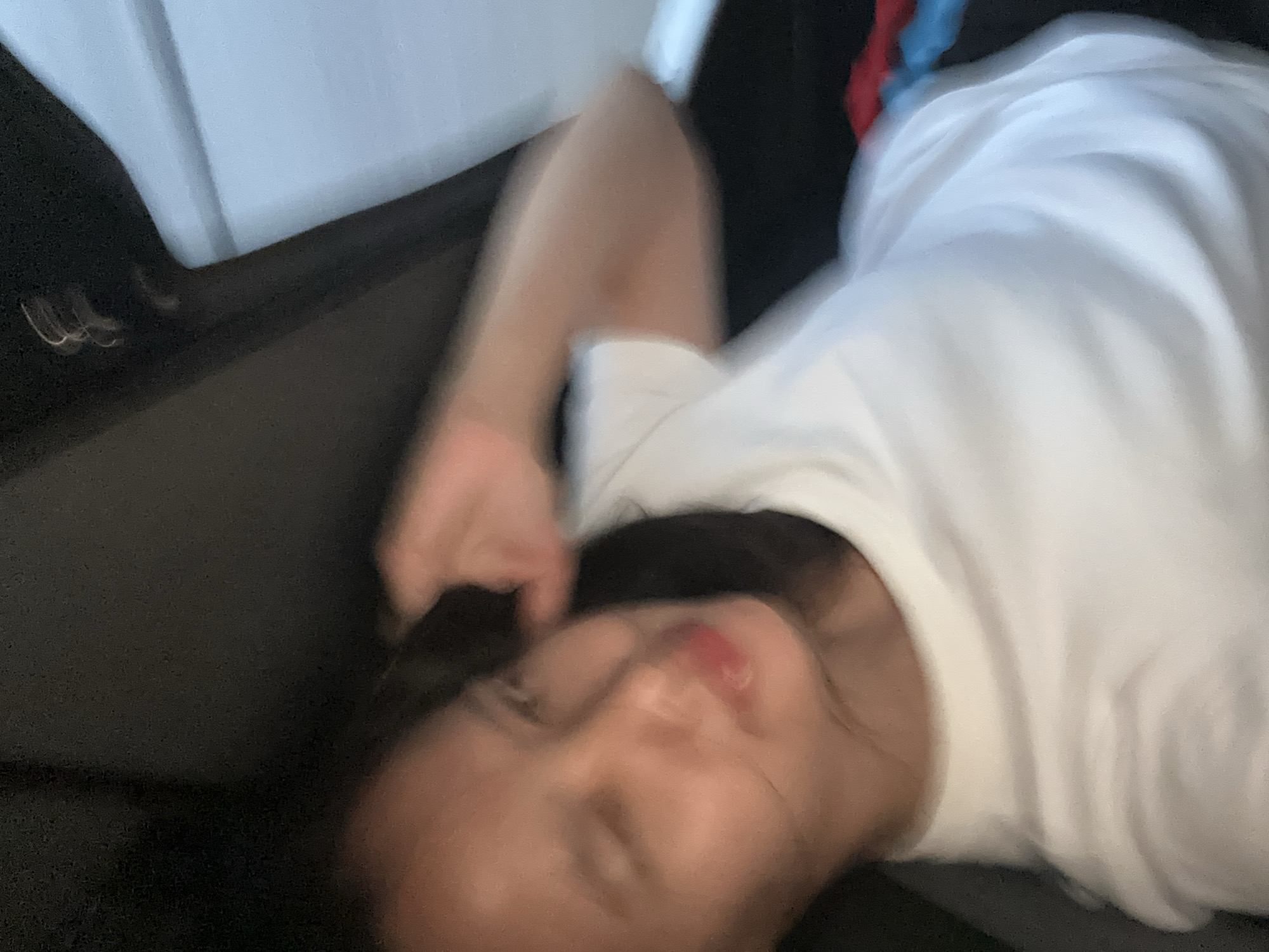 The image shows a close-up, slightly blurred, indoor shot of a young woman wearing a white t-shirt. She appears to be in a dimly lit environment, possibly a vehicle or a room with limited light. Her right hand is raised towards her head, obscuring part of her face, and her eyes are partially closed, giving the impression she is resting or posing for the photo. The background is dark with some indistinct objects visible on the right side, including what appears to be red and blue fabric. The image does not contain enough information to determine the specific location within Davao City, Philippines, or any urban context.
