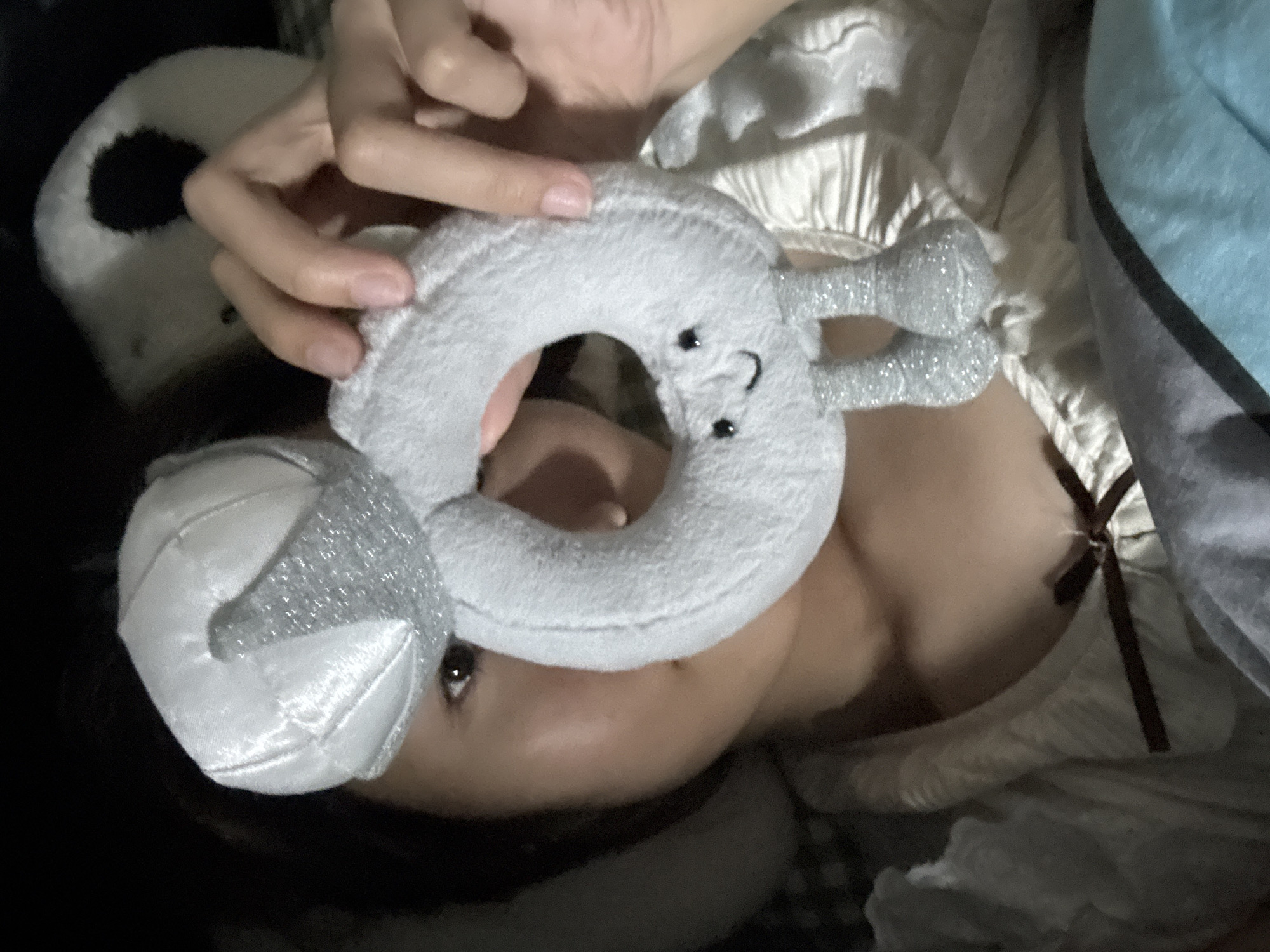 A person is lying down, possibly in bed, holding a plush toy up to the camera. The toy is a ring shape with a face and an attachment that resembles a gemstone. The person is wearing a light-colored top with a dark ribbon detail. In the background is a white plush object, possibly a pillow or stuffed animal, and what appears to be bedding or a blanket. The location of Nan'an District, China is not discernible from the visual information provided.