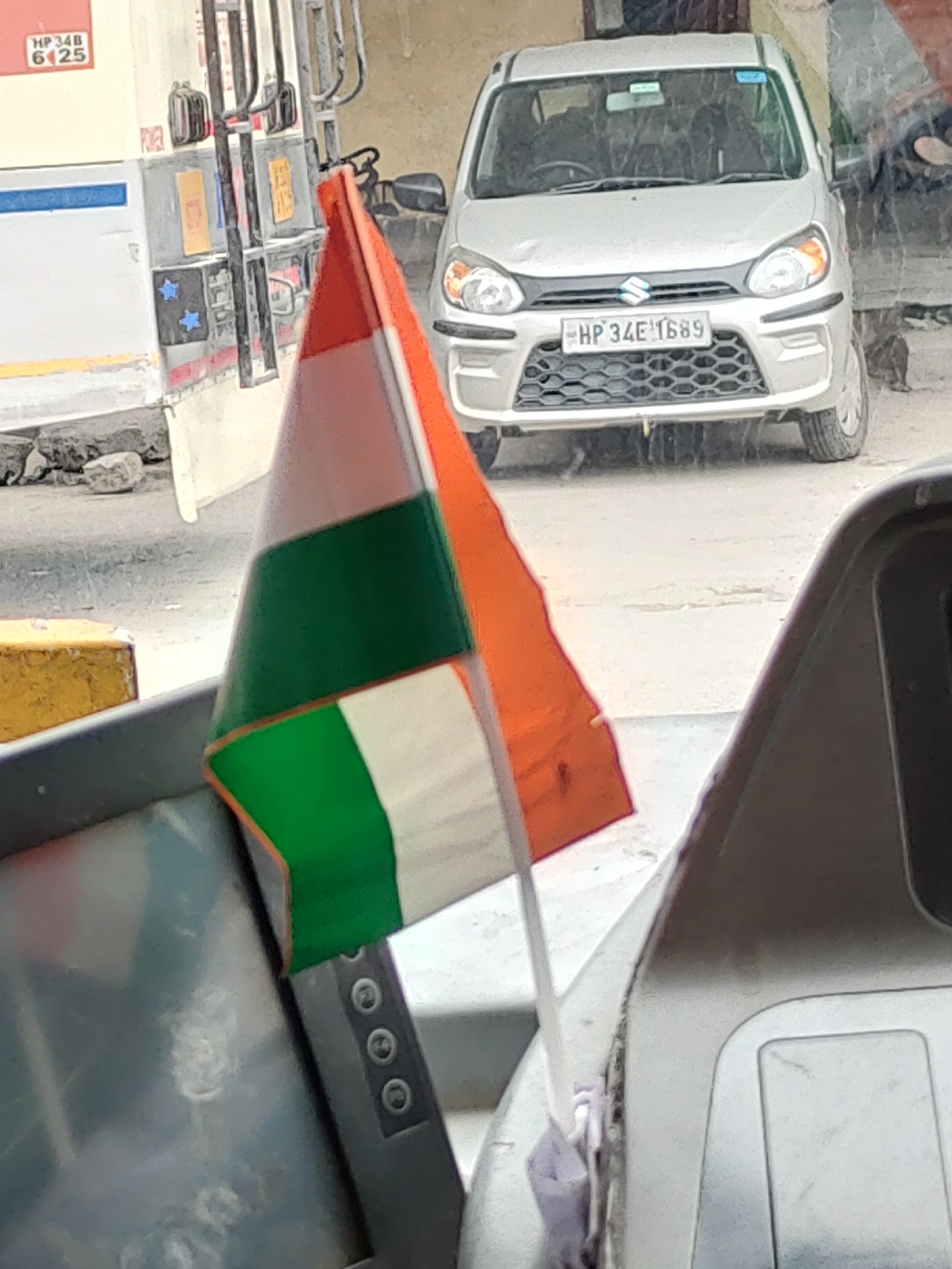 An Indian flag, orange, white, and green, is held by an unseen individual and is positioned in the foreground, obscuring parts of a gray car and a cream-colored vehicle. The car, a silver Maruti Suzuki Alto, is in the middle ground with its license plate, HP 34E 1689, visible. The cream-colored vehicle, which appears to be a bus or a large truck, is partially visible on the left. The background consists of a textured wall and what seems to be a street or pavement. The overall scene suggests an outdoor setting in Manali, India, possibly near a roadside establishment.