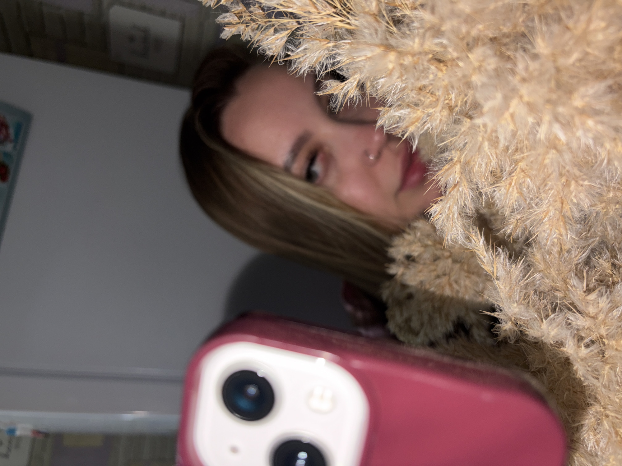 A woman is partially visible behind a bundle of dried wheat or similar grain stalks, taking a selfie with a pink phone. The background is out of focus, but appears to be an interior space. There is no information to place the image in Port-Katon, Russia.