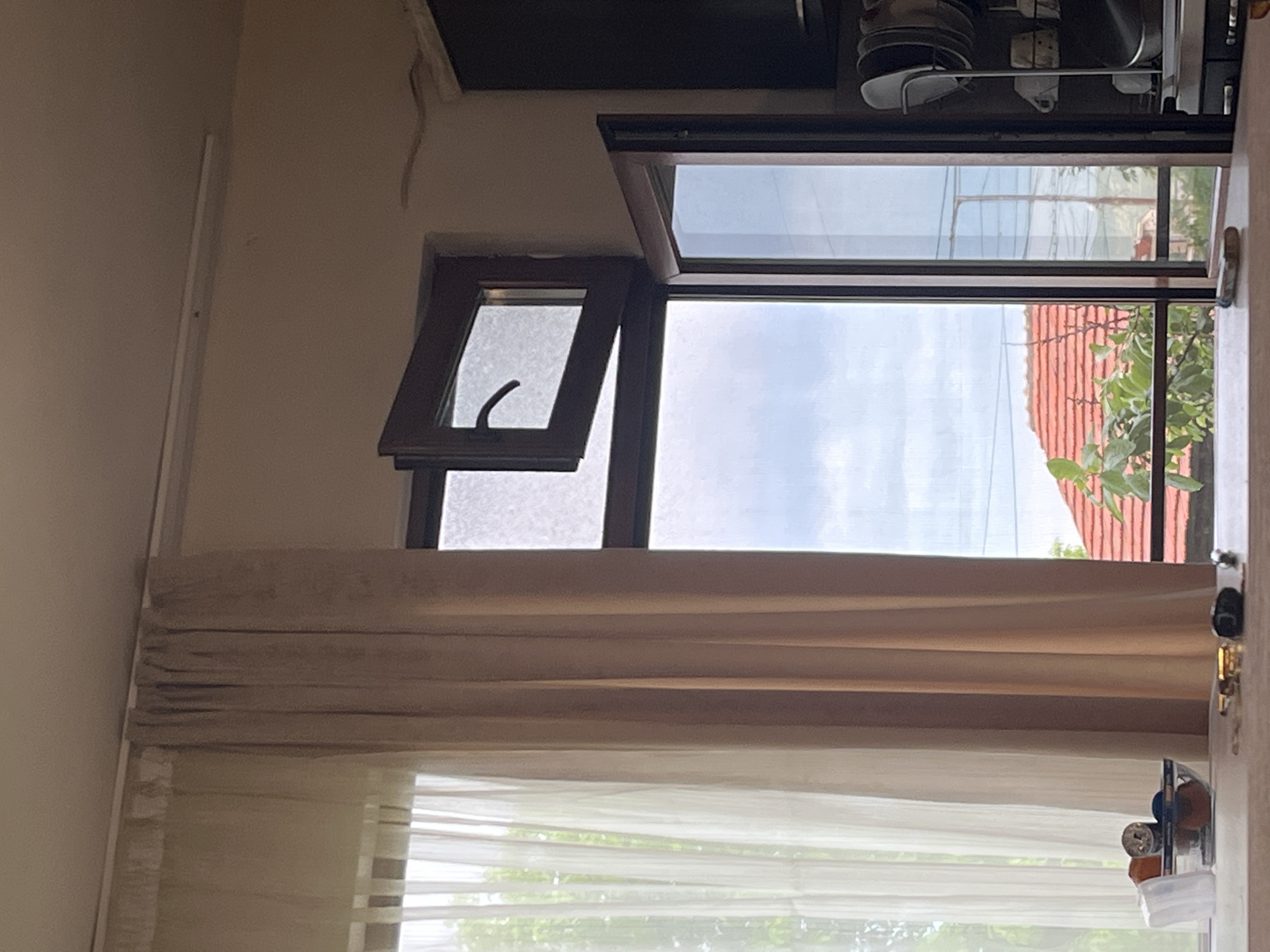 The image displays an indoor scene, primarily focused on a large window during daytime. The window features dark frames, with a small upper-left section angled open inwards. Covering the window are two layers: an outer, beige or light brown Roman blind partially drawn down, and a sheer white curtain behind it, covering the lower portion.
Above the window, on a dark, shelf-like structure, various items are visible, including stacked plates in a dish rack and other metallic objects, suggesting a kitchen or utility area. The interior walls are light-colored, and a white strip, possibly a light fixture or molding, runs along the left wall near the ceiling. To the right of the window, on a surface, a clear container holds small objects like bottles and a flashlight.
Through the window, a bright, partly cloudy sky is visible. Below the sky, a distinct red-tiled roof, partially obscured by green tree foliage, can be seen, hinting at an architectural style common in the region. Utility lines or wires are also visible against the sky in the upper outdoor section.
No people or specific activities are present. The bright light indicates it is daytime, and the outdoor view, particularly the red-tiled roof, is consistent with the provided location context of Burgas, Bulgaria. There is no visible text.