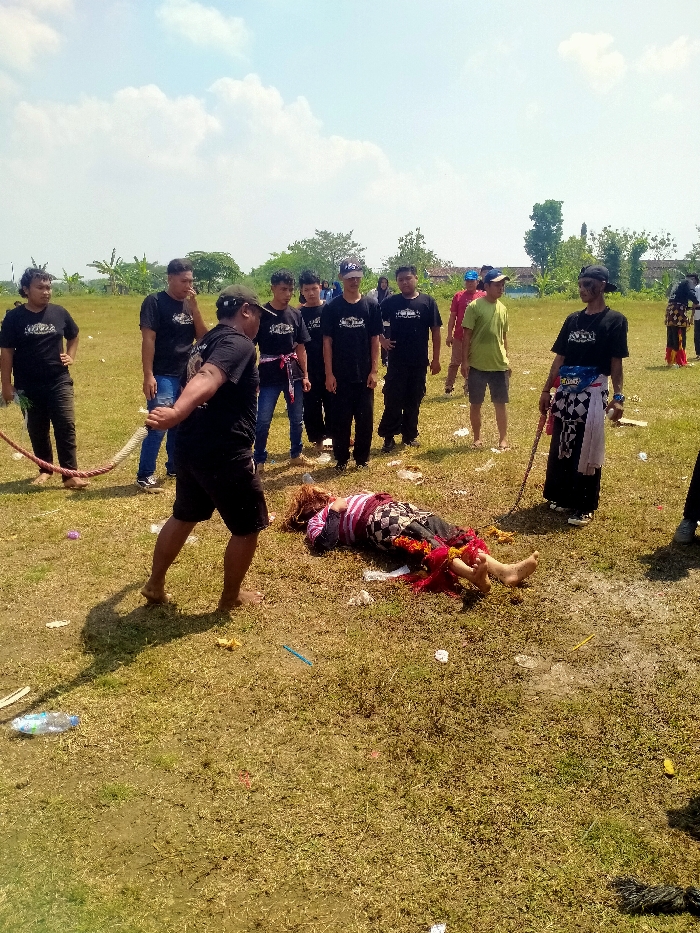 This image depicts an outdoor scene in Celep, Indonesia, on a clear, sunny day in a dry, grassy field.
**Environment:** The setting is an open field with sparse, dry grass and patches of exposed soil. The ground is littered with various discarded items, including plastic bottles, wrappers, and colored debris. In the background, scattered trees, shrubs, and indistinct distant structures are visible under a bright blue sky with sparse white clouds. Strong sunlight creates distinct shadows to the right of the standing figures.
**People and Subjects:** Approximately 12 individuals are present.
* **Central Figure:** A person of indeterminate gender lies prone (face down) on the grass in the center-right foreground. The individual wears a horizontally striped top (dark, pink, white), a dark patterned lower garment (possibly a sarong or skirt), and red fabric with tassels wrapped around the lower legs/ankles. Their feet are bare. The head is turned to the left, and arms appear bent beneath the body.
* **Foreground Left Participant:** An adult male, barefoot and wearing a dark t-shirt and dark shorts, is positioned in the immediate foreground with his back partially to the viewer. He is holding a thick, braided light-colored rope that extends out of frame to the left.
* **Mid-Ground Left Group:** A group of approximately seven adult males stands observing, primarily wearing dark t-shirts (some with logos), some with caps, and some with colorful sashes or belts. One individual in this group also holds the rope.
* **Mid-Ground Right Individuals:** Two adult males are present. One wears a lime green t-shirt and light shorts. The other wears a dark t-shirt, a dark cap, and dark sunglasses, with a checkered black and white sash/skirt over dark pants. This individual holds a thin, dark stick vertically.
* **Background Right Figure:** A partially visible individual in the far right background also appears to be wearing traditional attire, including red fabric, similar to the central prone figure.
**Actions and Interactions:** The central figure is motionless in a prone position on the ground. The surrounding individuals are standing and observing the scene. The rope on the left suggests an ongoing or recently concluded activity, possibly a tug-of-war or ritualistic performance, with multiple participants holding it. No direct physical interaction with the prone individual is evident at the moment captured.