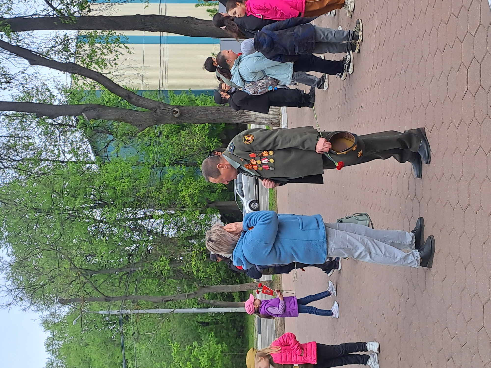 A line of people stands in a park-like area in Elektrostal, Russia. The ground is paved with light brown hexagonal bricks. A man in a green military uniform with multiple medals and a red flower in his left hand stands in the line. He is carrying a military hat. Behind the man, a row of adults and children stand with their heads tilted back, seemingly looking at something in the sky. A woman in a blue jacket is visible with her right hand to her forehead. Several children are also in the line, one in a pink jacket, another in a purple jacket holding red flowers. A building with white and blue panels is visible in the background behind the trees. A silver car is also in the background. Green trees are present on the left and behind the subjects.
