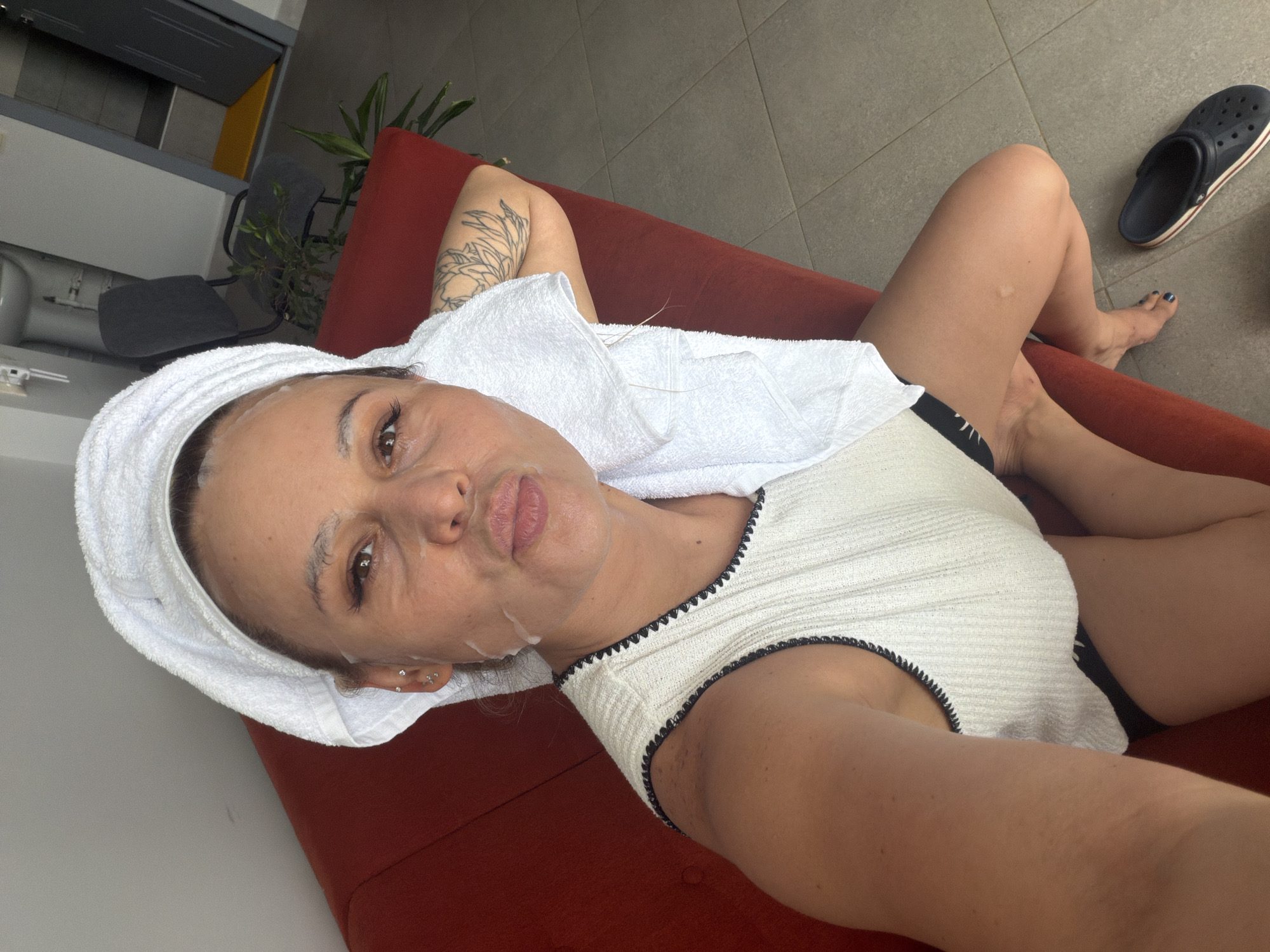 A woman with a facial mask and a white towel wrapped around her head is sitting on a red couch. She is wearing a white sleeveless top with black trim and black underwear. She has a tattoo on her left arm. A white towel is draped around her neck and shoulders. She is looking at the camera with her lips puckered. To the right of the couch, a black Croc shoe is on the floor. The environment appears to be indoors, possibly in an apartment, with tiled flooring visible. In the background, a desk chair, a plant, a doorway, and a toilet can be seen, suggesting it might be a living space or a bathroom area. The location is specified as Sarapul, Russia.
