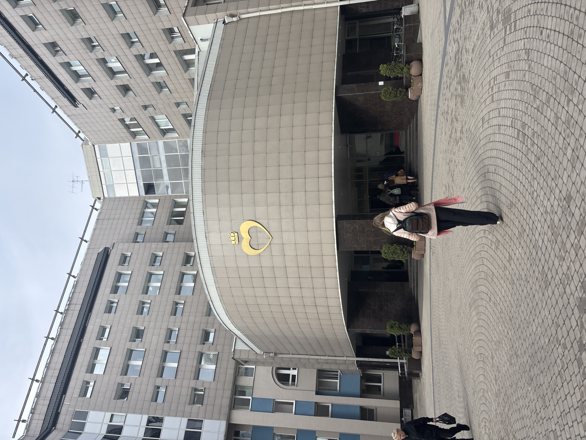 The image depicts the building of the Herzen University in Saint Petersburg, Russia. The building is modern, with a curved facade and gray tiled exterior. A gold emblem of a heart and crown is visible on the facade. People are visible in the foreground and near the entrance of the building. The ground is paved with gray cobblestones in a circular pattern. The sky is overcast.
