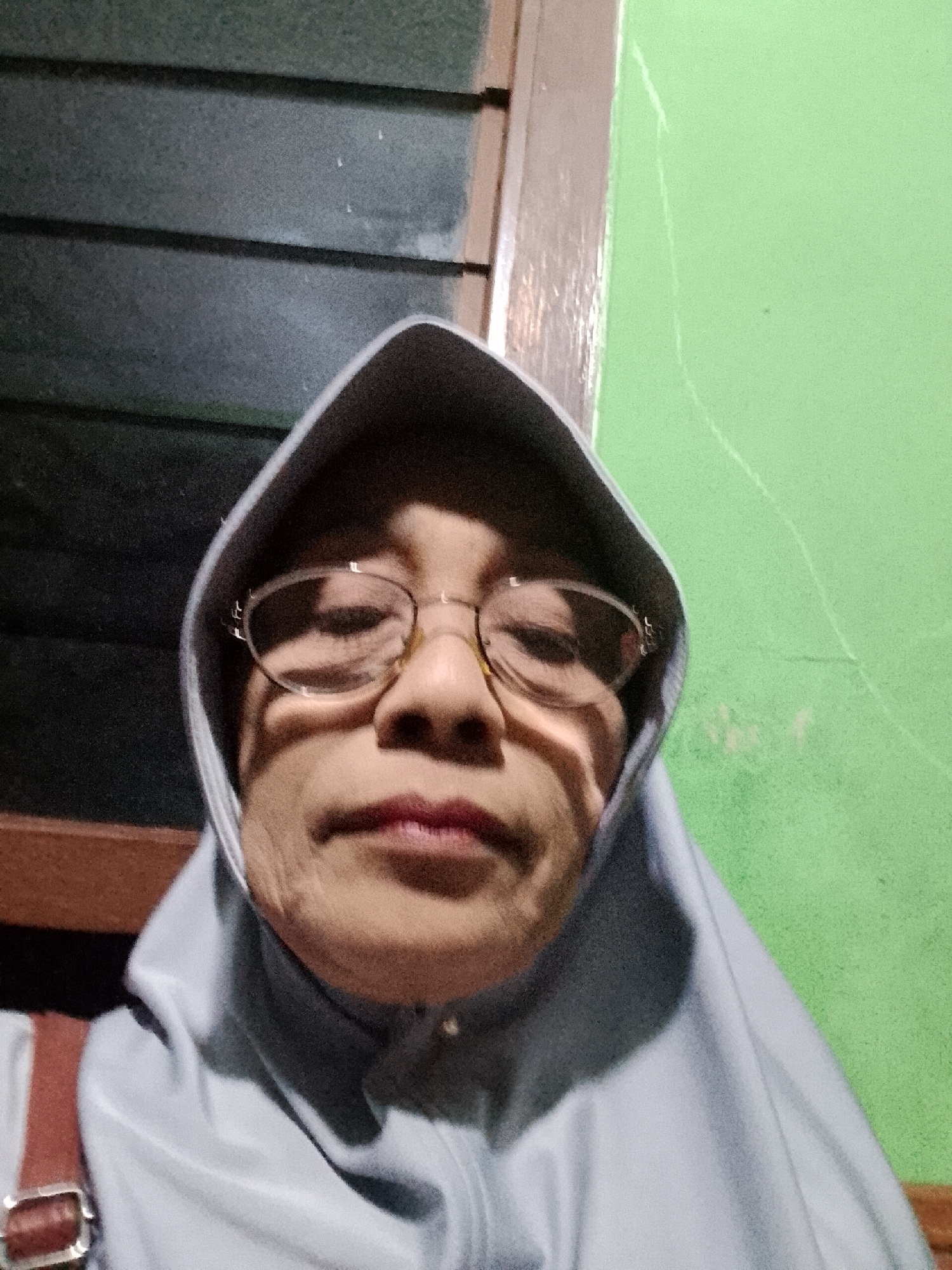 The image is a selfie of a woman wearing glasses and a grey hijab. She is indoors, with a dark, slatted surface behind her left shoulder and a green wall behind her right. The lighting suggests it is evening or indoors with artificial light. There is no indication of her location within Kecamatan Pati, Indonesia. The woman's face is the primary focus, and the background elements provide context of an indoor setting.